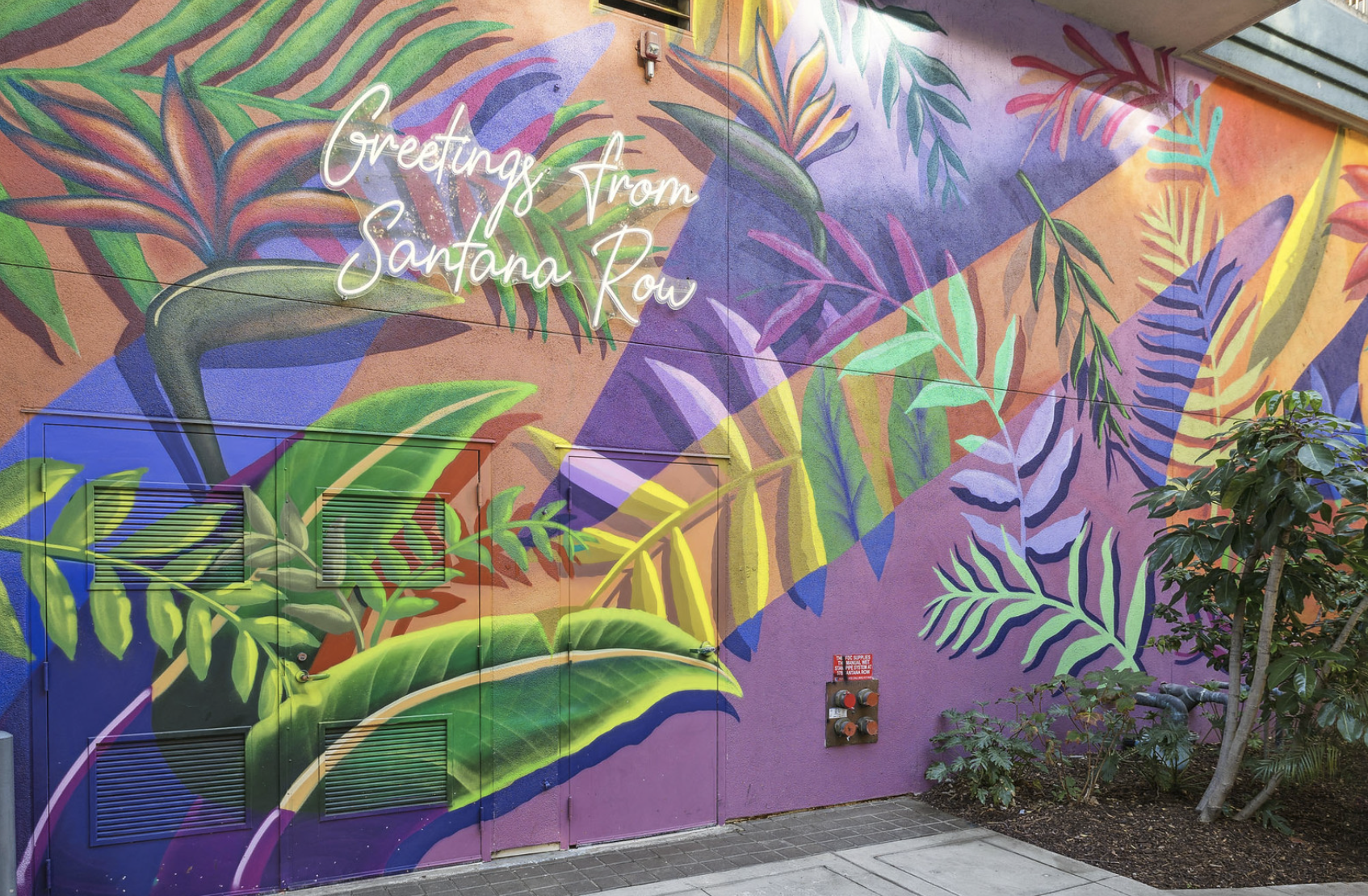 Colorful mural with tropical leaves and plants on a pink and purple wall. White neon-style text reads 'Greetings from Santana Row.' There is a tree and some plants in the foreground, and utility vents and pipes on the wall.