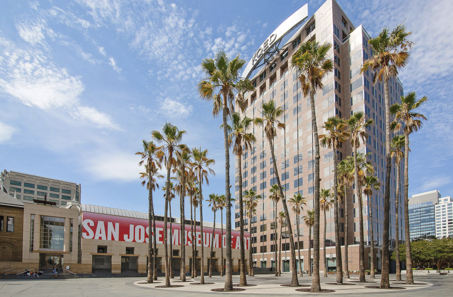 Tall modern building with the sign 'TOWER' on the top, surrounded by a row of palm trees, with a smaller building labeled 'SAN JOSE MUSEUM OF ART' in the foreground on a sunny day.