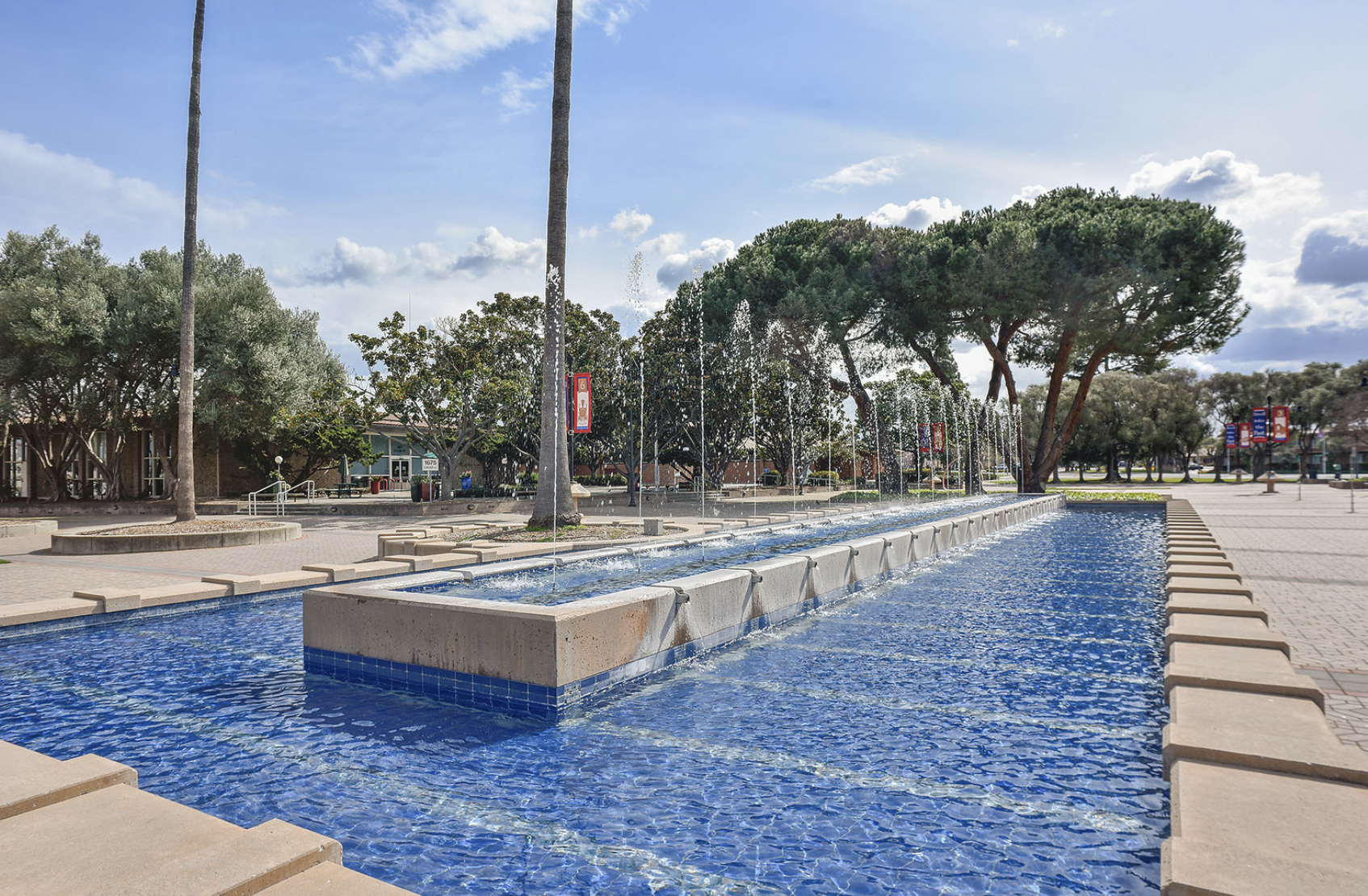 A wide view of an outdoor plaza with a long rectangular water fountain, tall palm trees, other trees, and banners hanging on posts. The sky is partly cloudy and sunny.