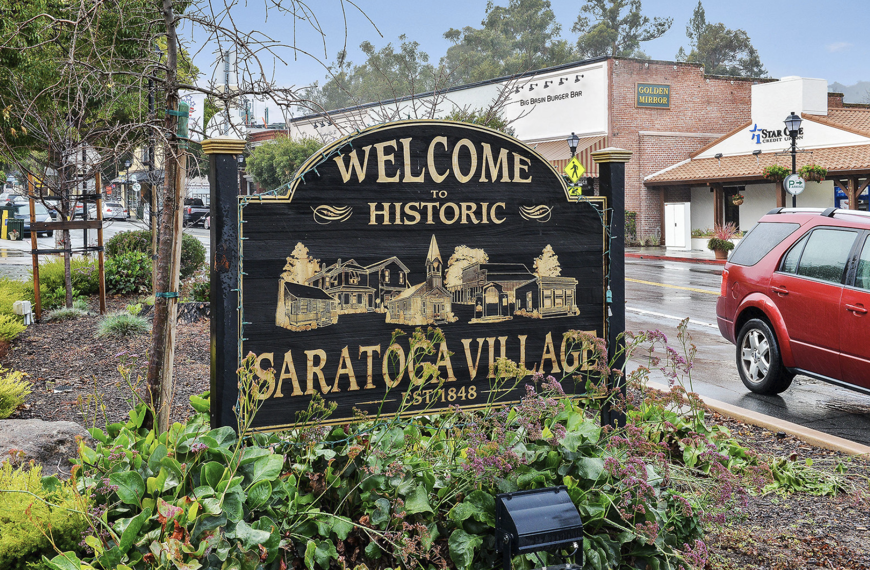 A black sign with gold lettering welcomes visitors to Saratoga Village, established in 1848, featuring illustrations of historic buildings, situated on a street with a parked red vehicle and storefronts in the background.