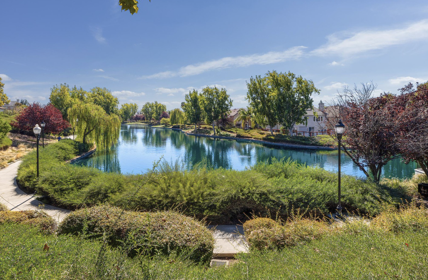 A peaceful park scene with a calm river surrounded by green, purple, and red trees, a paved walkway, and vintage-style street lamps under a bright blue sky.