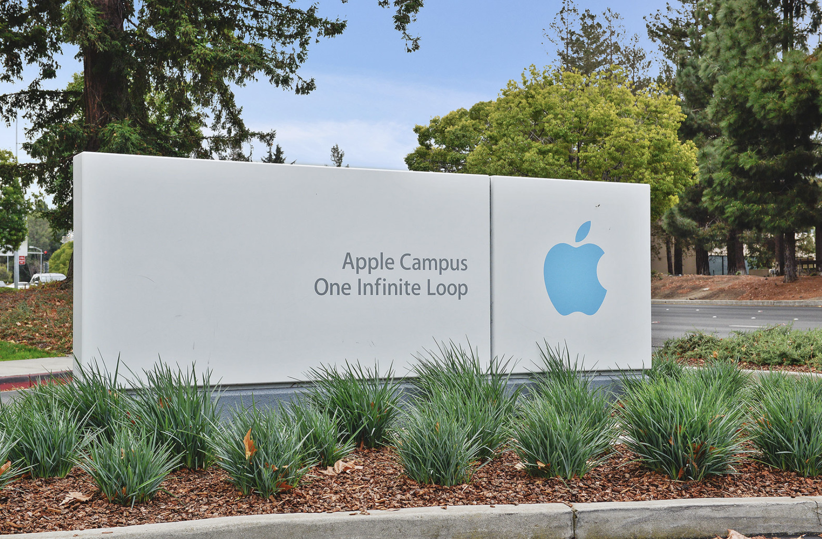 Large white sign with blue Apple logo and text that reads 'Apple Campus One Infinite Loop' in front of green plants, trees, and a street.