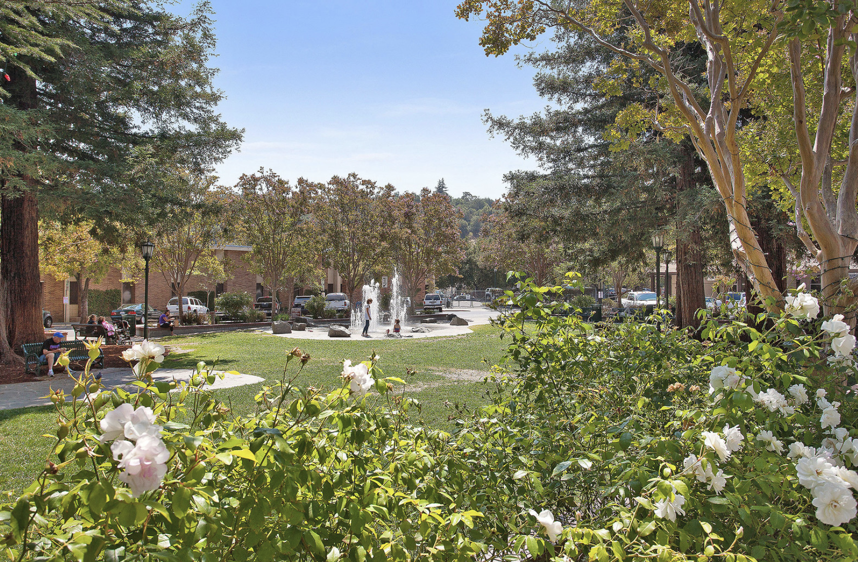 A park with a fountain, surrounded by trees and flowers, with people sitting on benches and children playing.