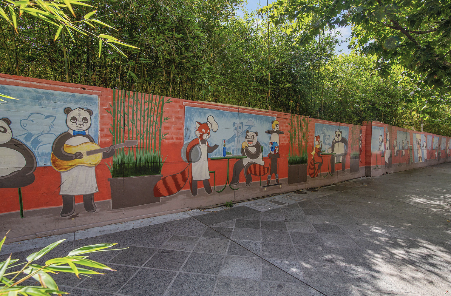 Colorful mural featuring anthropomorphic pandas, foxes, and birds enjoying activities like playing guitar, dining, and socializing, painted on a long wall surrounded by plants and trees.