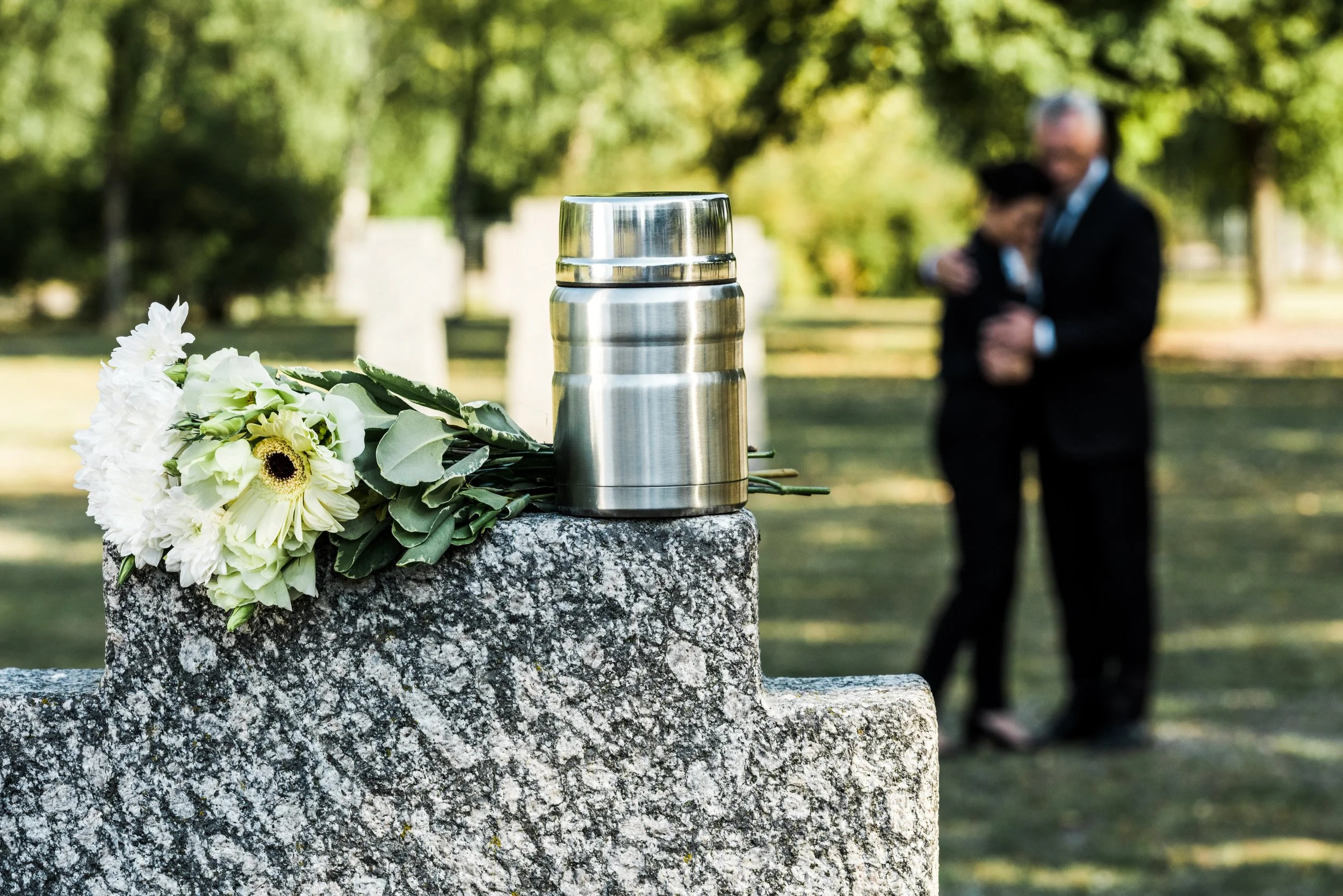 A small urn on a stone monument with a white floral wreath beside it, in a graveyard with trees and blurred figures in the background.