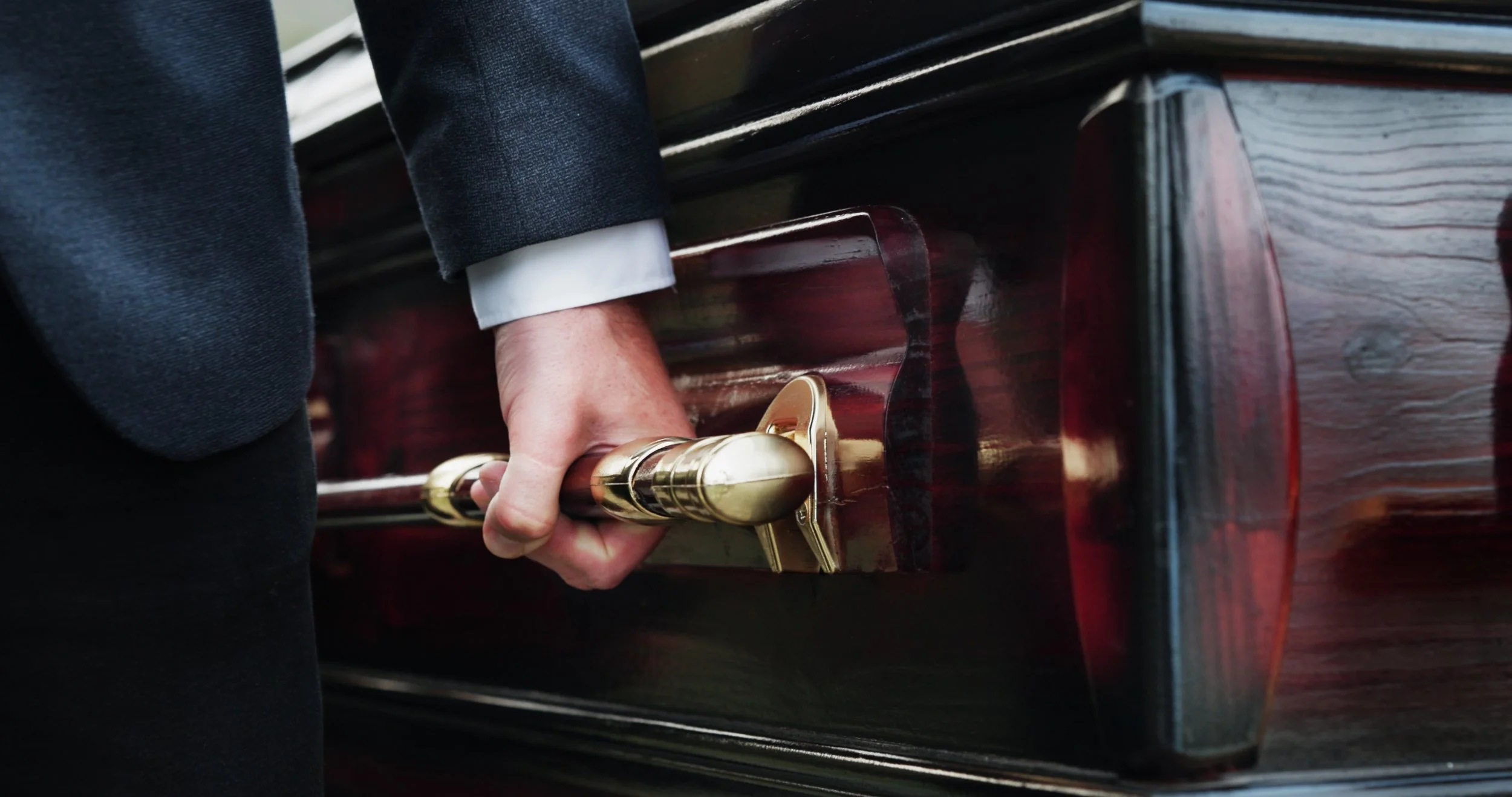 Close-up of a person's hand pressing the pedal of a piano, with the person wearing a dark suit and white shirt, and the piano being polished dark wood.