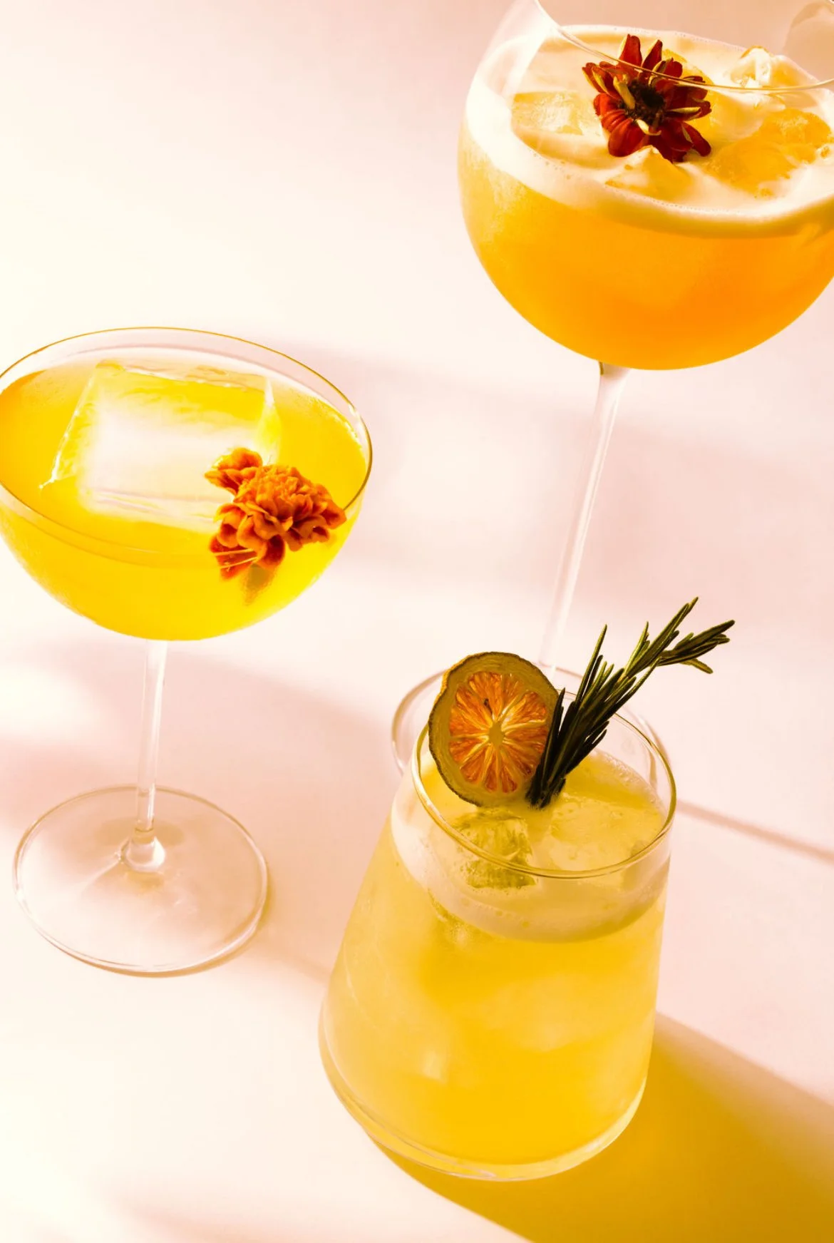 Three yellow cocktails with ice and floral garnishes, one in a coupe glass and two in highball glasses, with a lemon slice and rosemary sprig in one, all set against a light background.