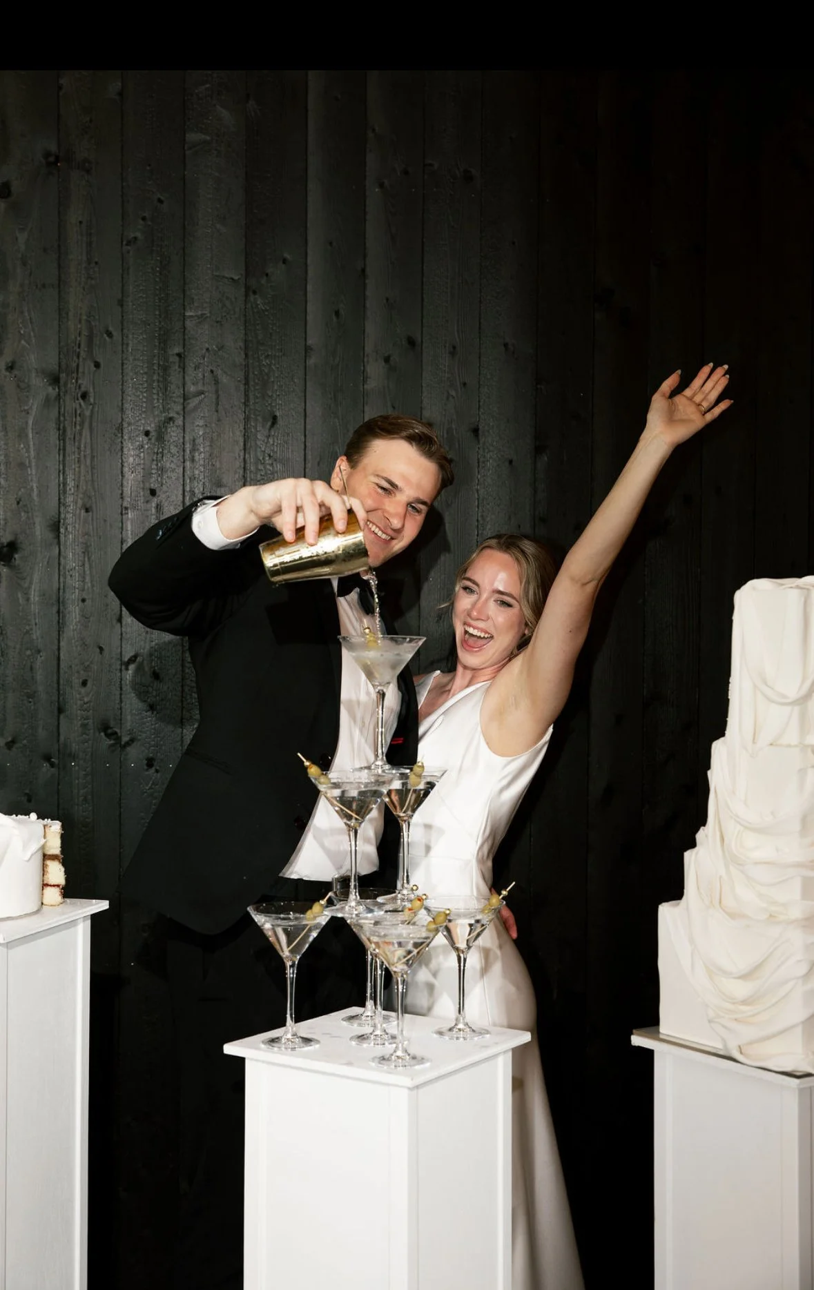 A man and woman celebrate at a wedding reception, pouring champagne into a tower of glasses. The woman is raising her arm in excitement. There are wedding cakes visible on the sides.