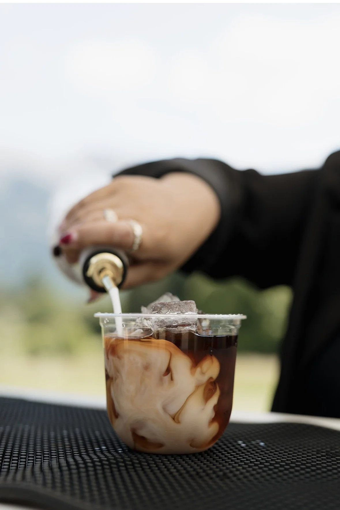 A person pouring cream into a glass of iced coffee with ice cubes, creating a swirl of cream and coffee on a black textured surface outdoors.