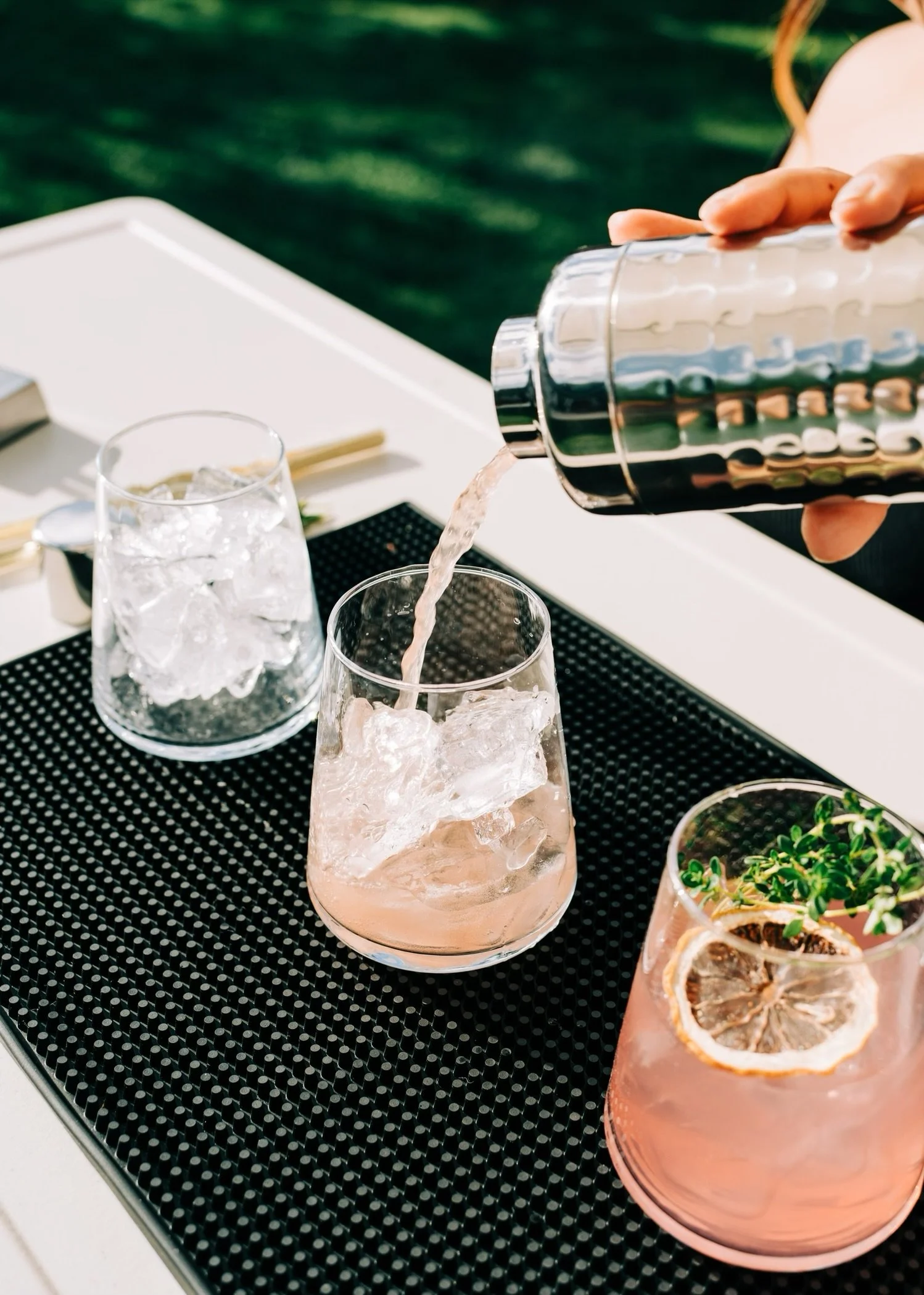 A person pouring a pink drink with ice into a glass on a black textured surface at an outdoor setting with greenery in the background.