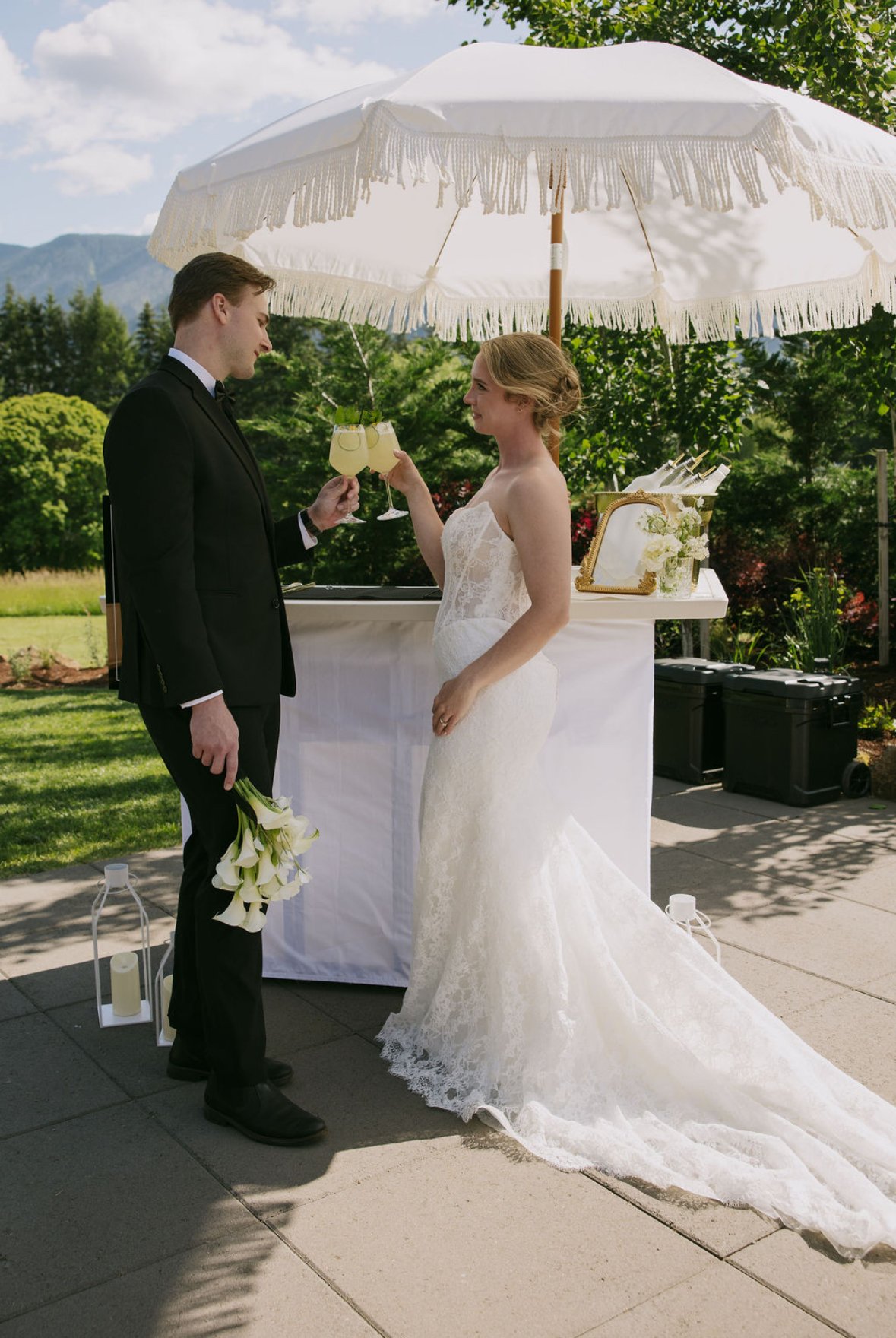 A bride and groom sharing a toast at their outdoor wedding ceremony. The groom is holding a bouquet of white flowers, and the bride is wearing a white lace wedding dress. They are standing under a white patio umbrella with a fringed edge, with a garden and mountains in the background.