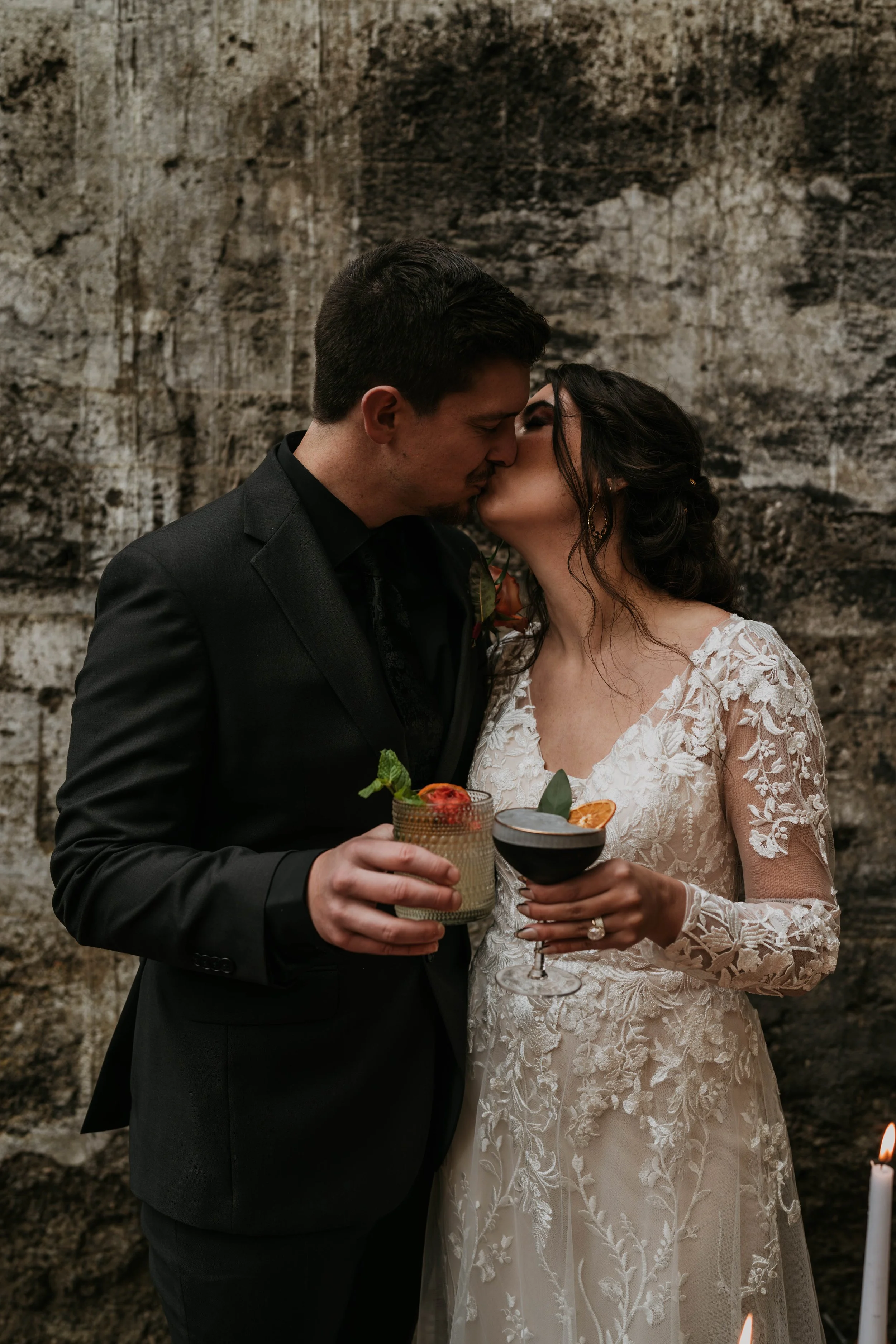 A couple dressed in wedding attire sharing a kiss at their wedding reception while holding cocktails.