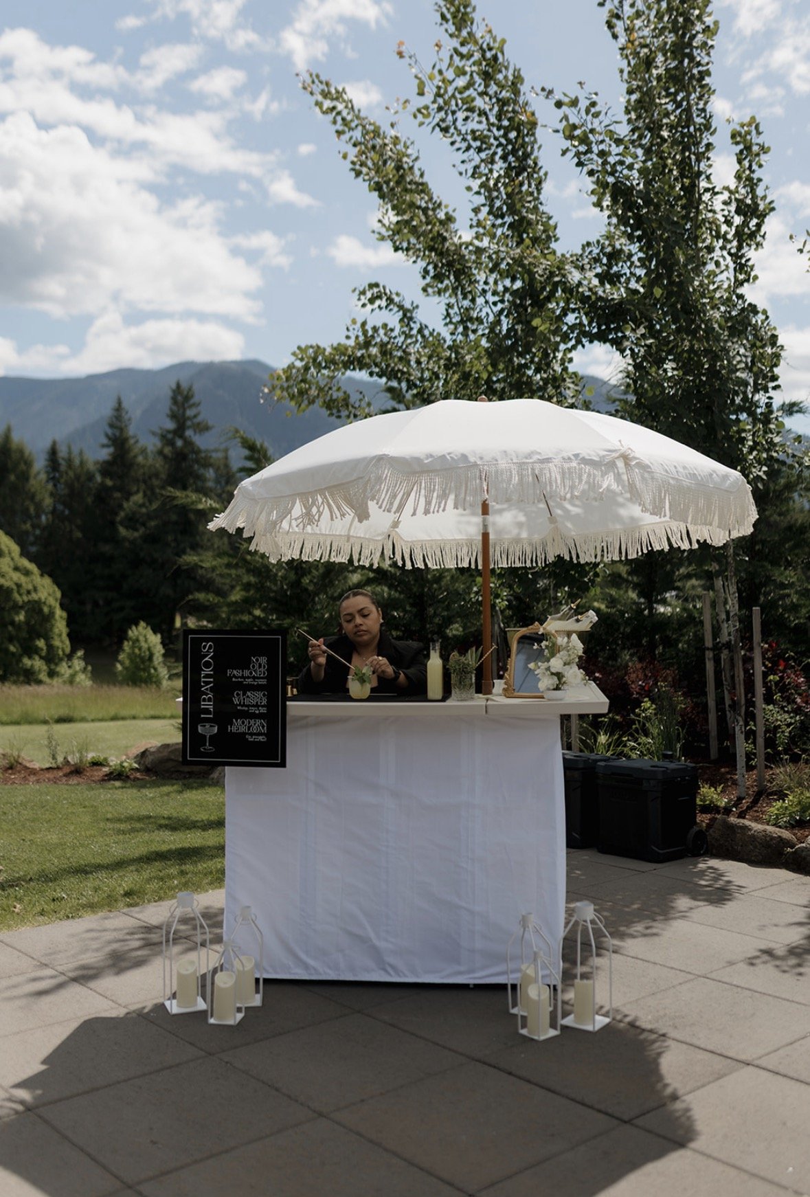 A woman stands behind a white outdoor bar under a large white umbrella, preparing a drink with mountain and tree scenery in the background. The bar is decorated with candles and flowers, and a sign advertising various cocktails is placed on the left.