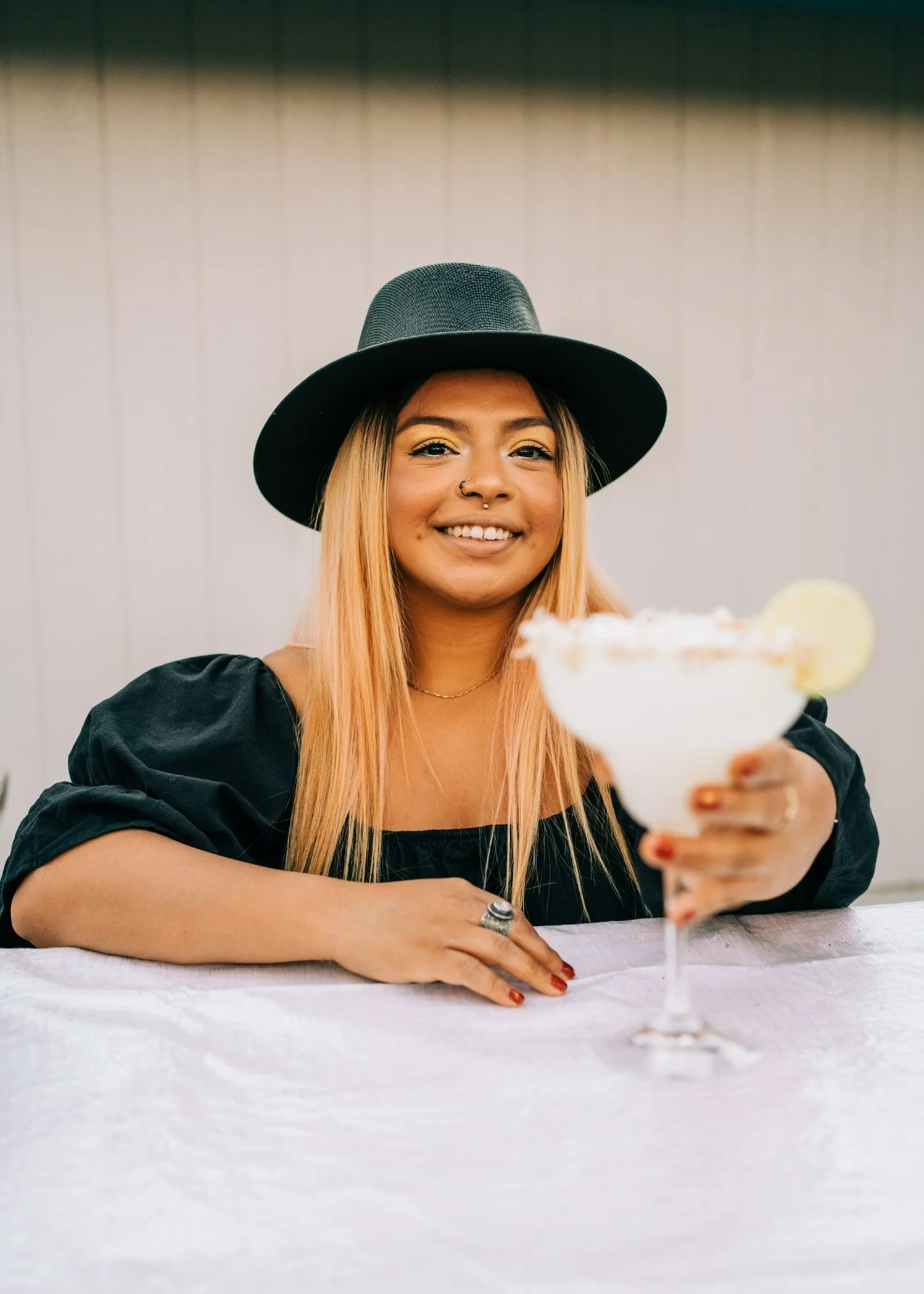 A woman with long blond hair, wearing a black hat and black dress, smiling and holding a margarita glass with lime garnish, sitting at a table with a white tablecloth.