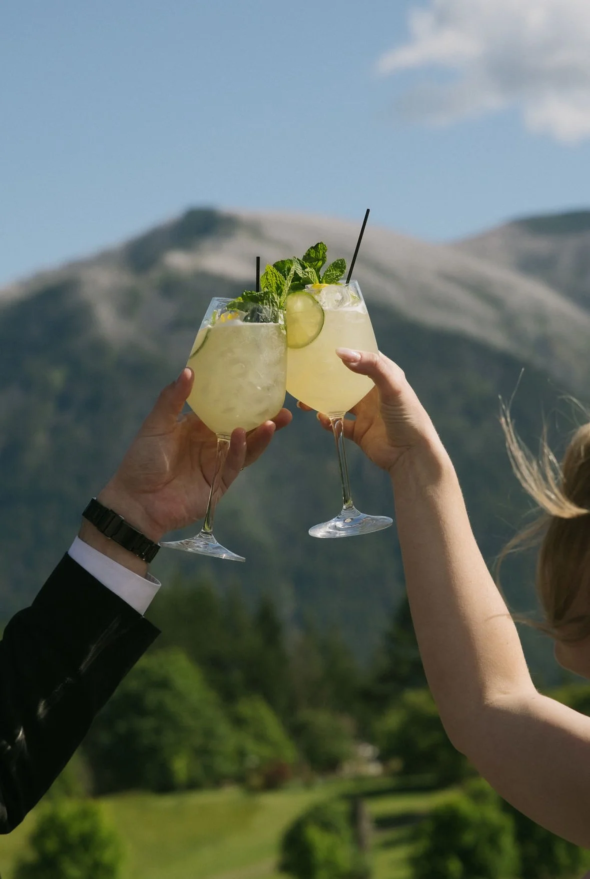 Two people raising glasses of yellow cocktail garnished with mint and cucumber slices outdoors with mountain background.