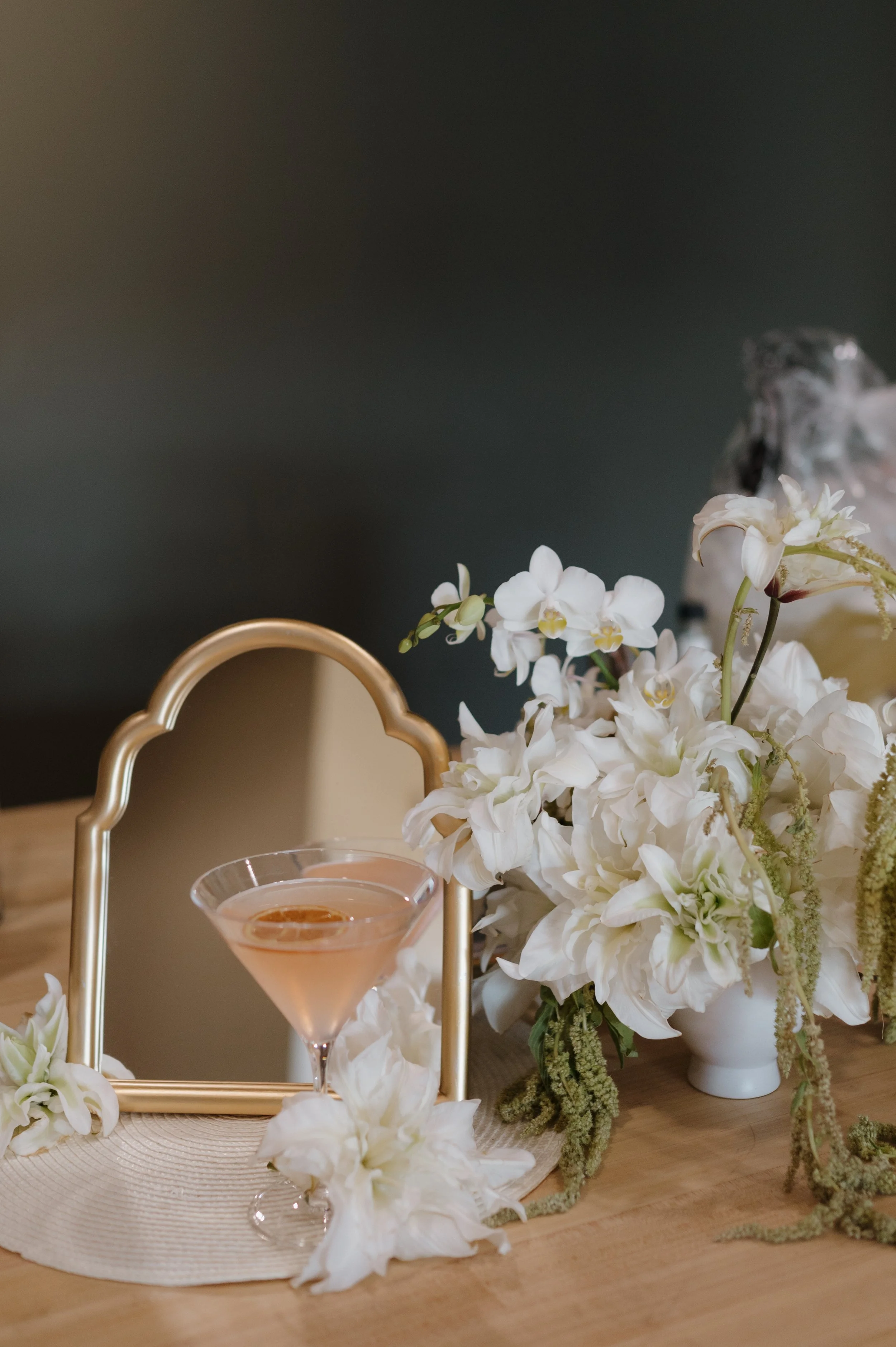 A table setting featuring a gold frame mirror, white flowers in a white vase, and a pink cocktail in a martini glass on a woven placemat.