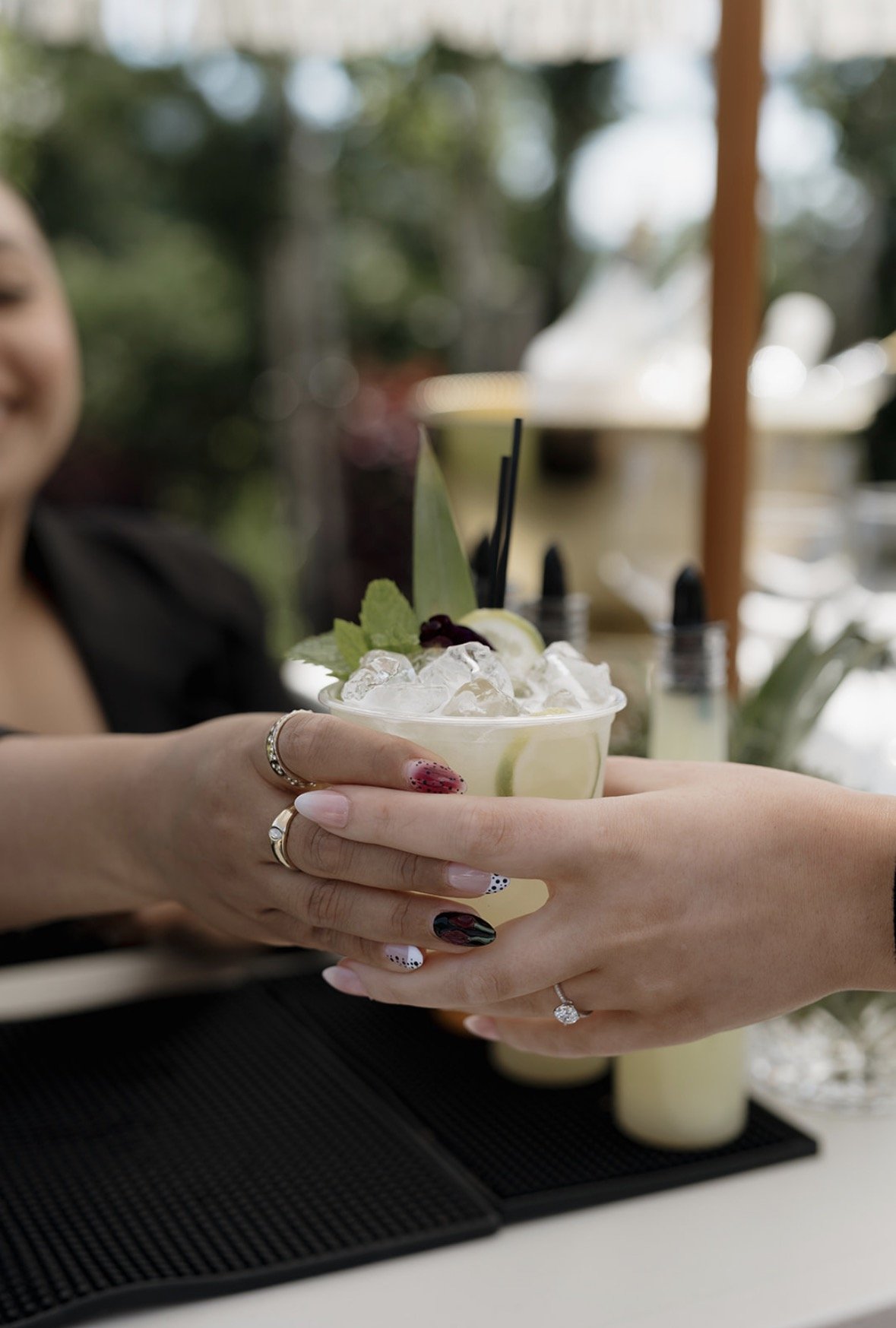 Two women exchanging a cocktail with ice, lime, and garnishes in a social setting. One woman has manicured nails and rings, and they are smiling.
