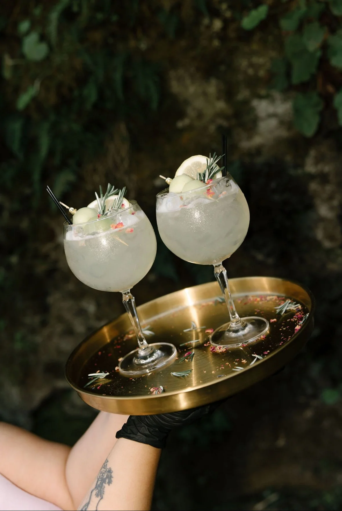 Two cocktail glasses with clear drinks garnished with lemon slices and herbs on a gold tray with flower petals, held by a person with a tattooed arm.