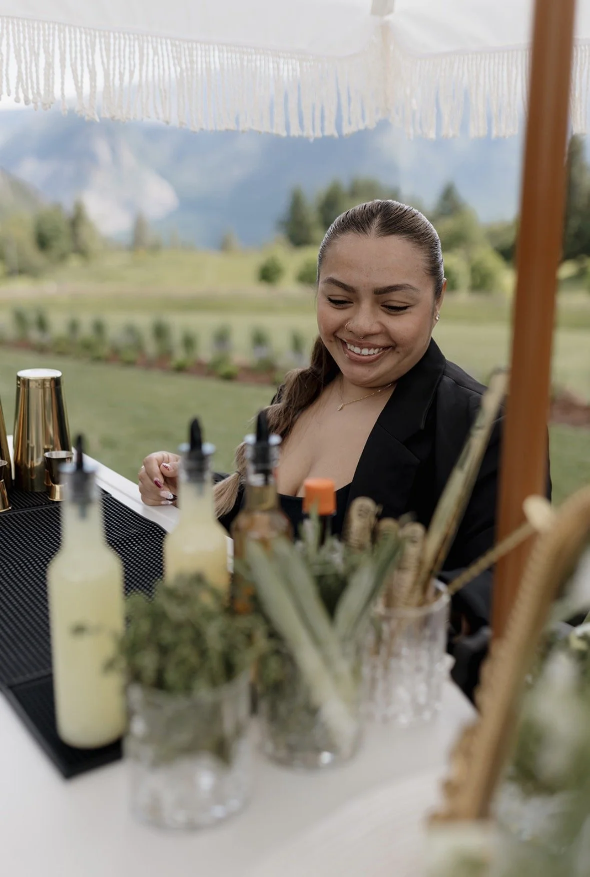 A woman with brown hair tied back, smiling while preparing drinks at an outdoor bar with bottles and bar tools, with a scenic mountain and green landscape in the background.