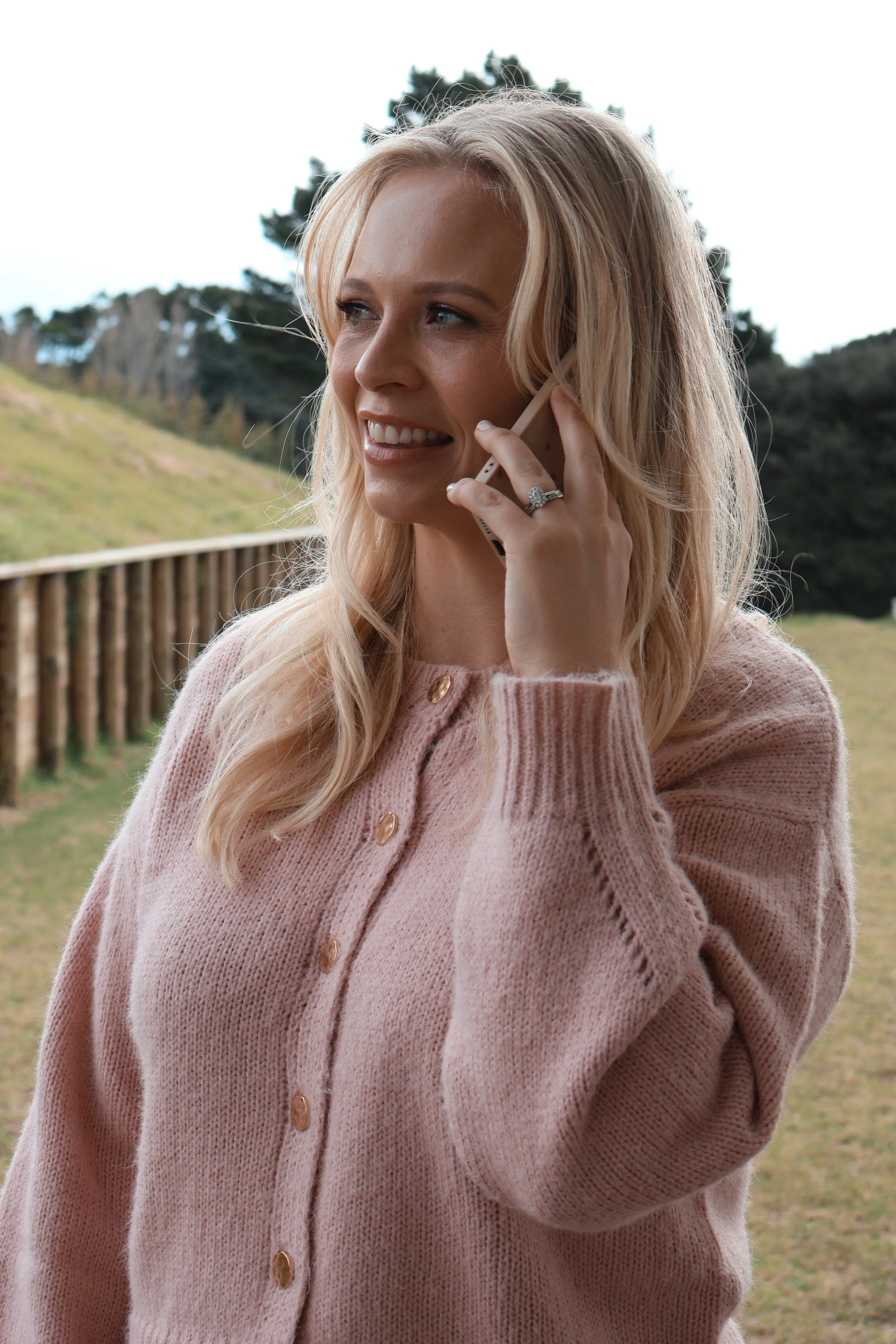 A woman with blonde hair smiling and talking on a mobile phone outdoors, wearing a pink knitted cardigan with wooden buttons, with trees and a grassy landscape in the background.