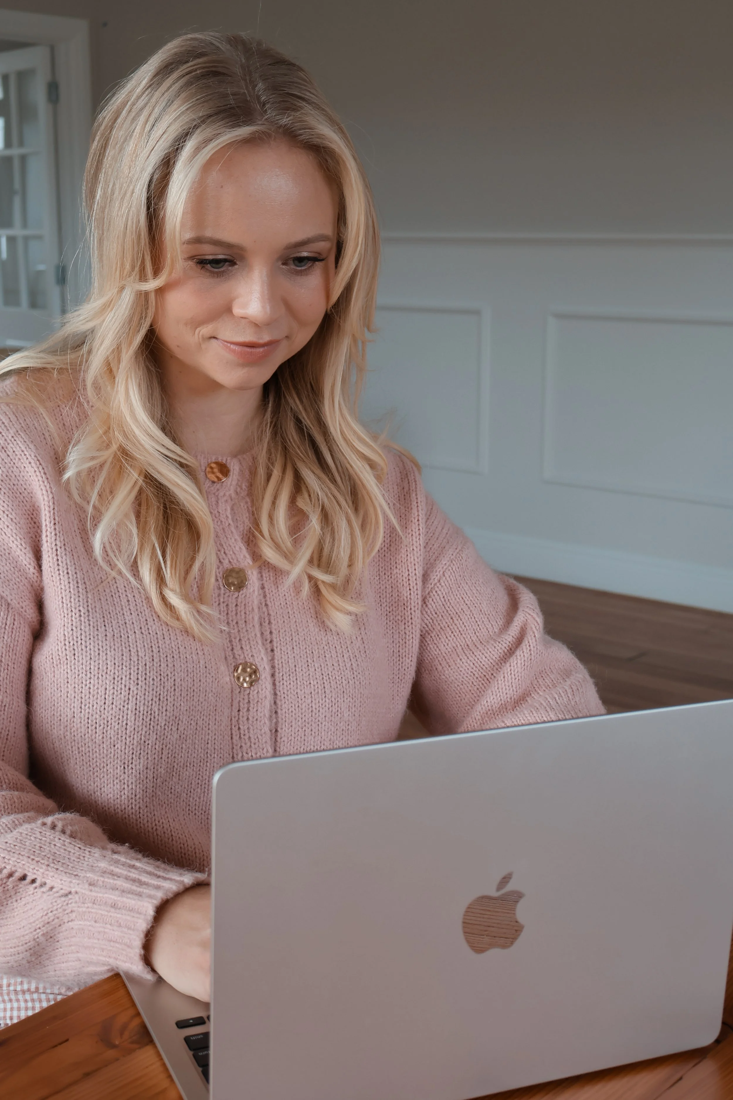 A woman with long blonde hair wearing a pink sweater sitting at a wooden table working on a silver MacBook laptop.