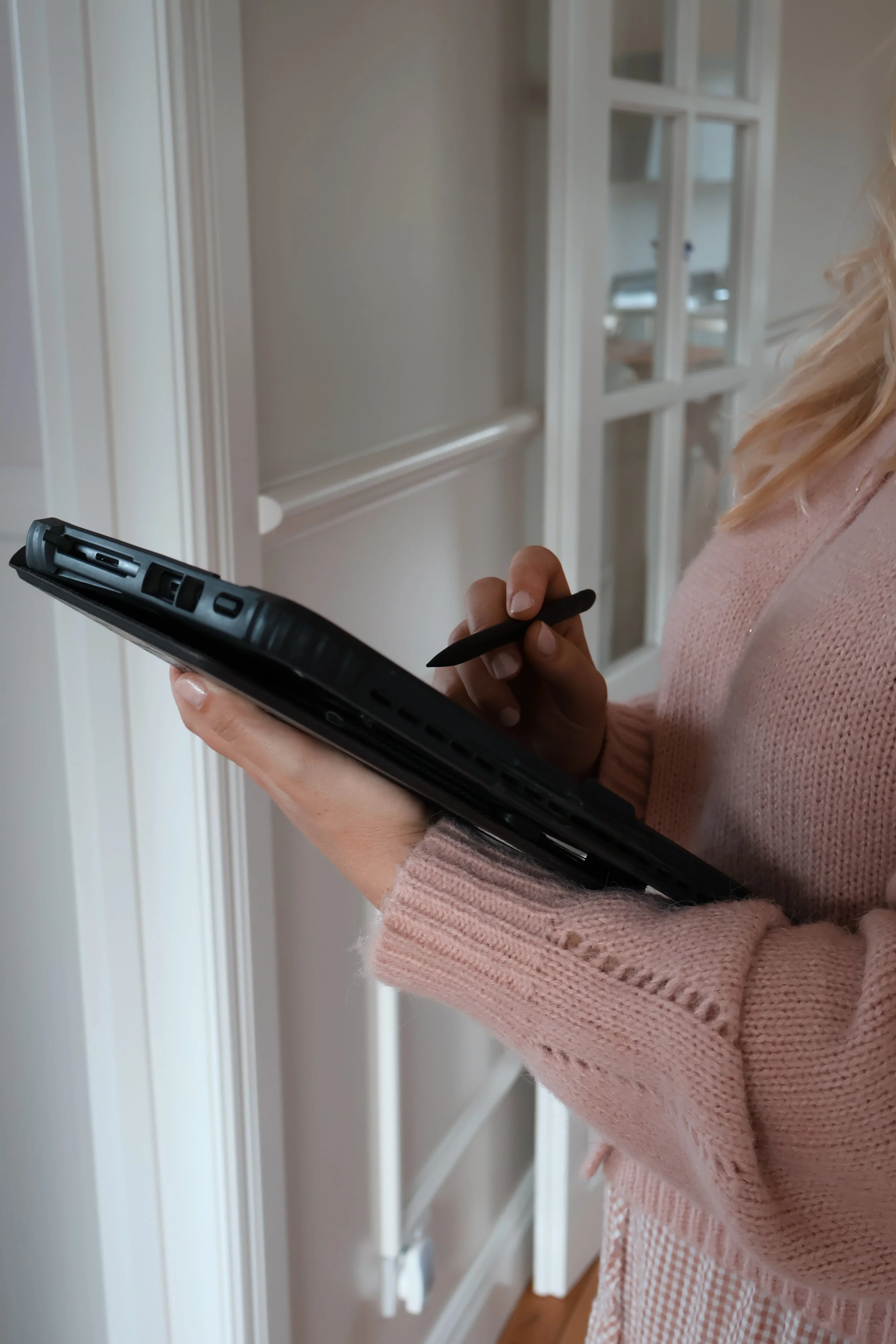 Close-up of a woman holding a tablet and stylus, wearing a pink sweater, standing in front of a white wall and a glass cabinet in a home interior.