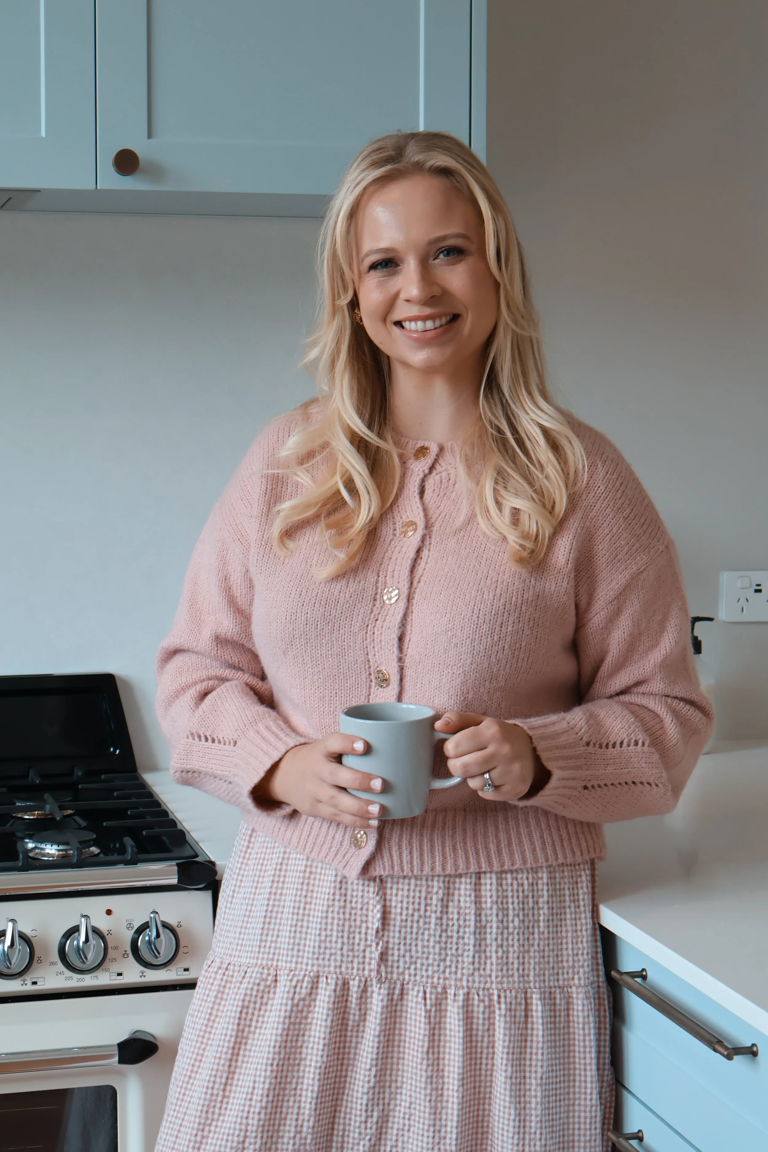 A woman with blonde hair and a pink cardigan is smiling and holding a mug while standing in a kitchen.