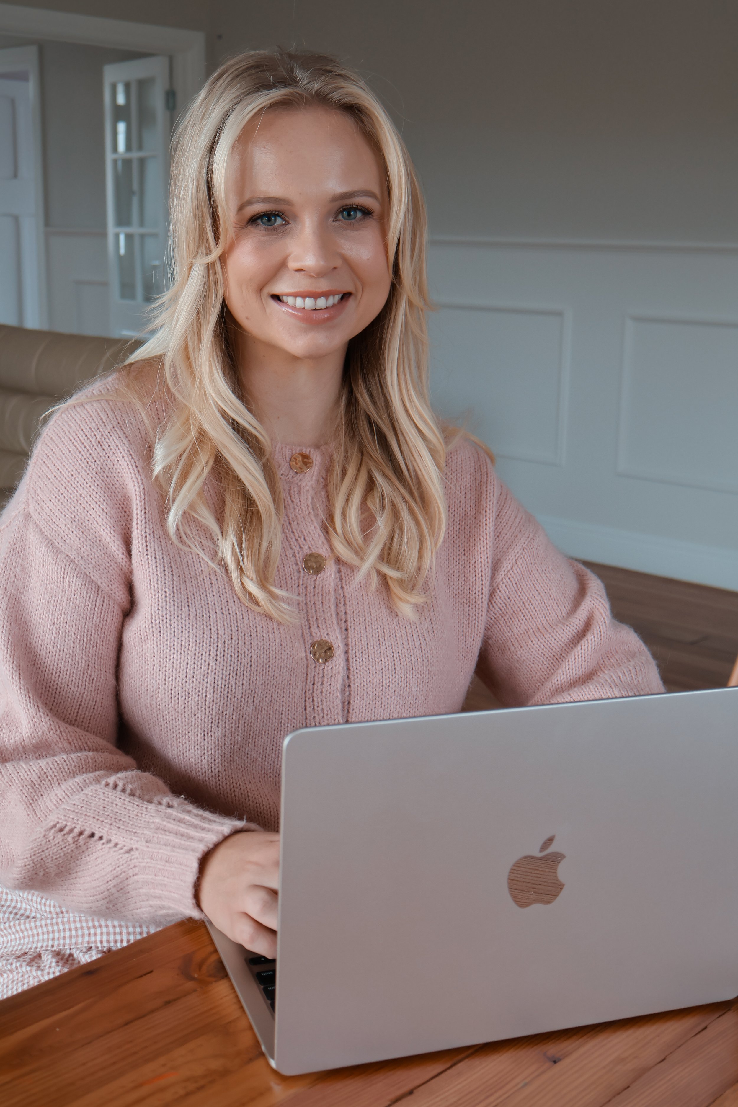 A blonde woman wearing a pink sweater is smiling while working on a silver MacBook laptop at a wooden table.