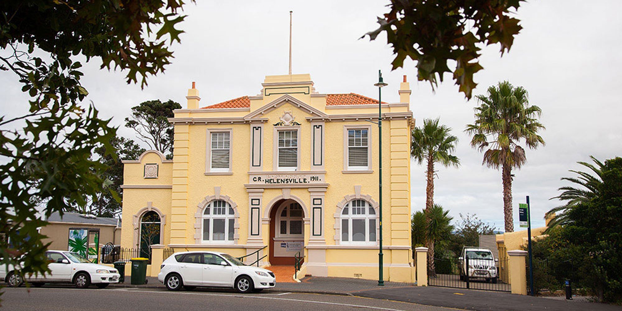 Yellow historic building with white trim, palm trees, parked cars, and cloudy sky.