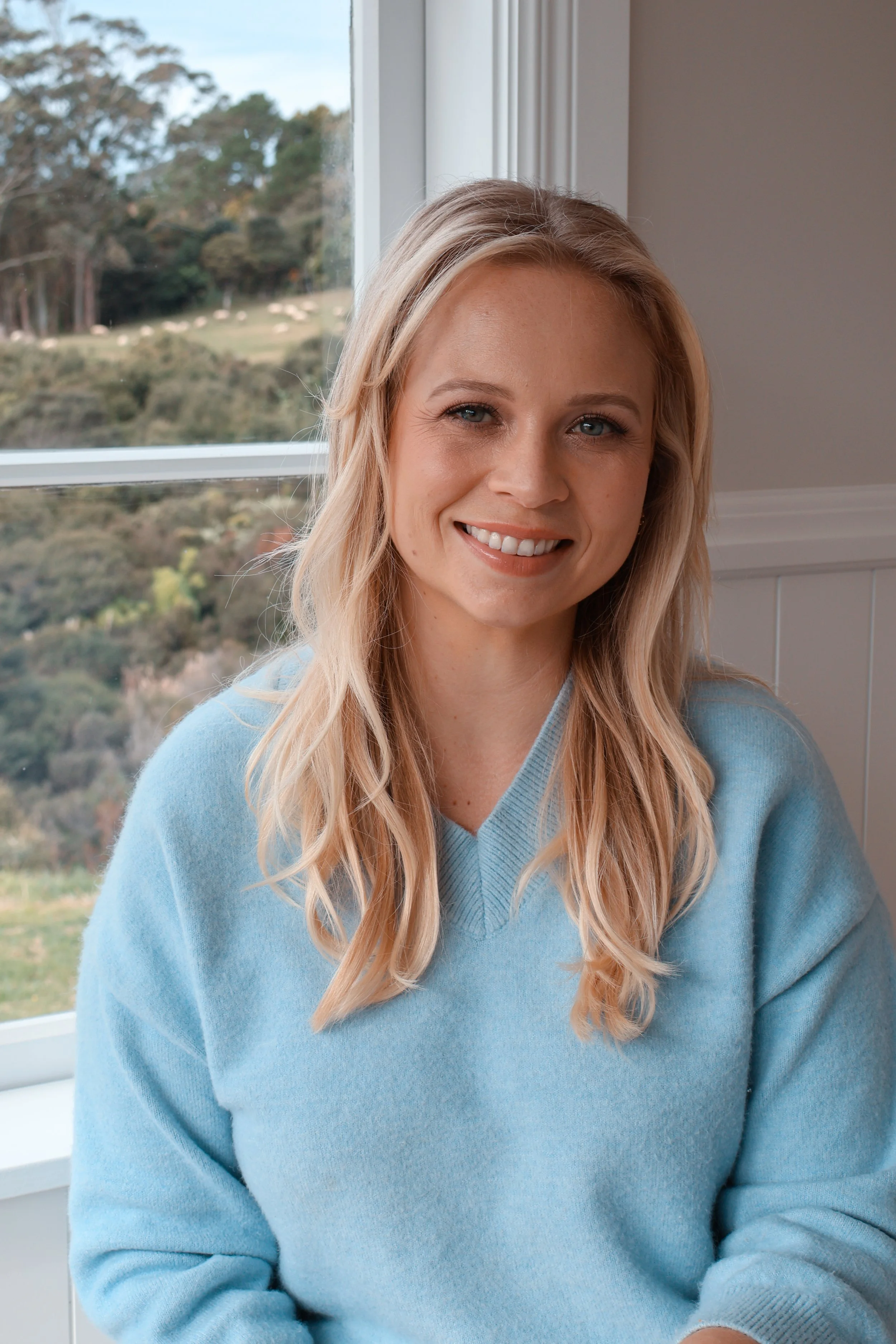 A woman with light blonde hair, wearing a light blue sweater, smiling near a window with a scenic outdoor view of trees and a hillside.