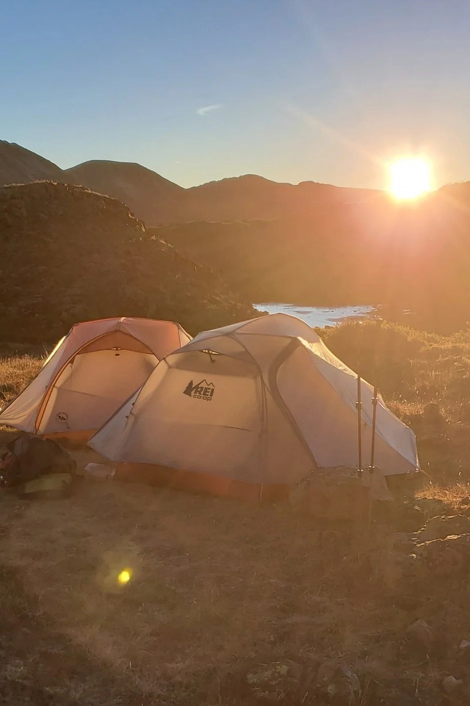 Two tents set up on a grassy hillside during sunset, with mountains, a lake, and a clear sky in the background.