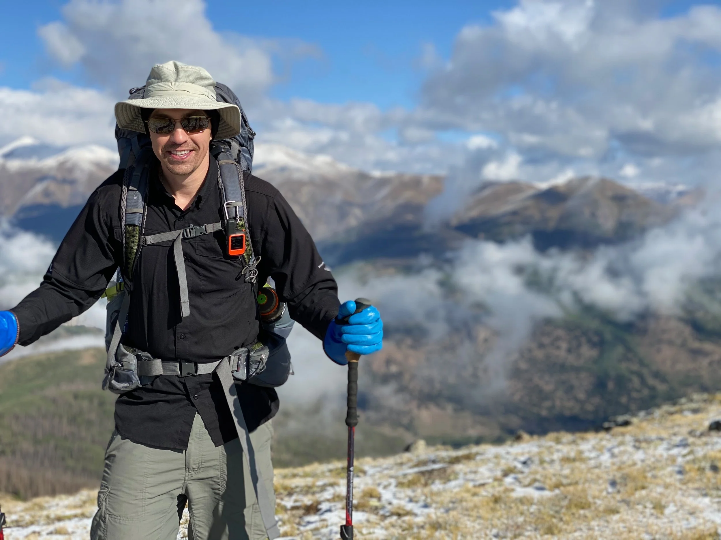 A smiling man dressed in outdoor hiking gear with a large backpack, hat, sunglasses, and hiking poles, standing on a mountain trail with snowy peaks and a partly cloudy sky in the background.