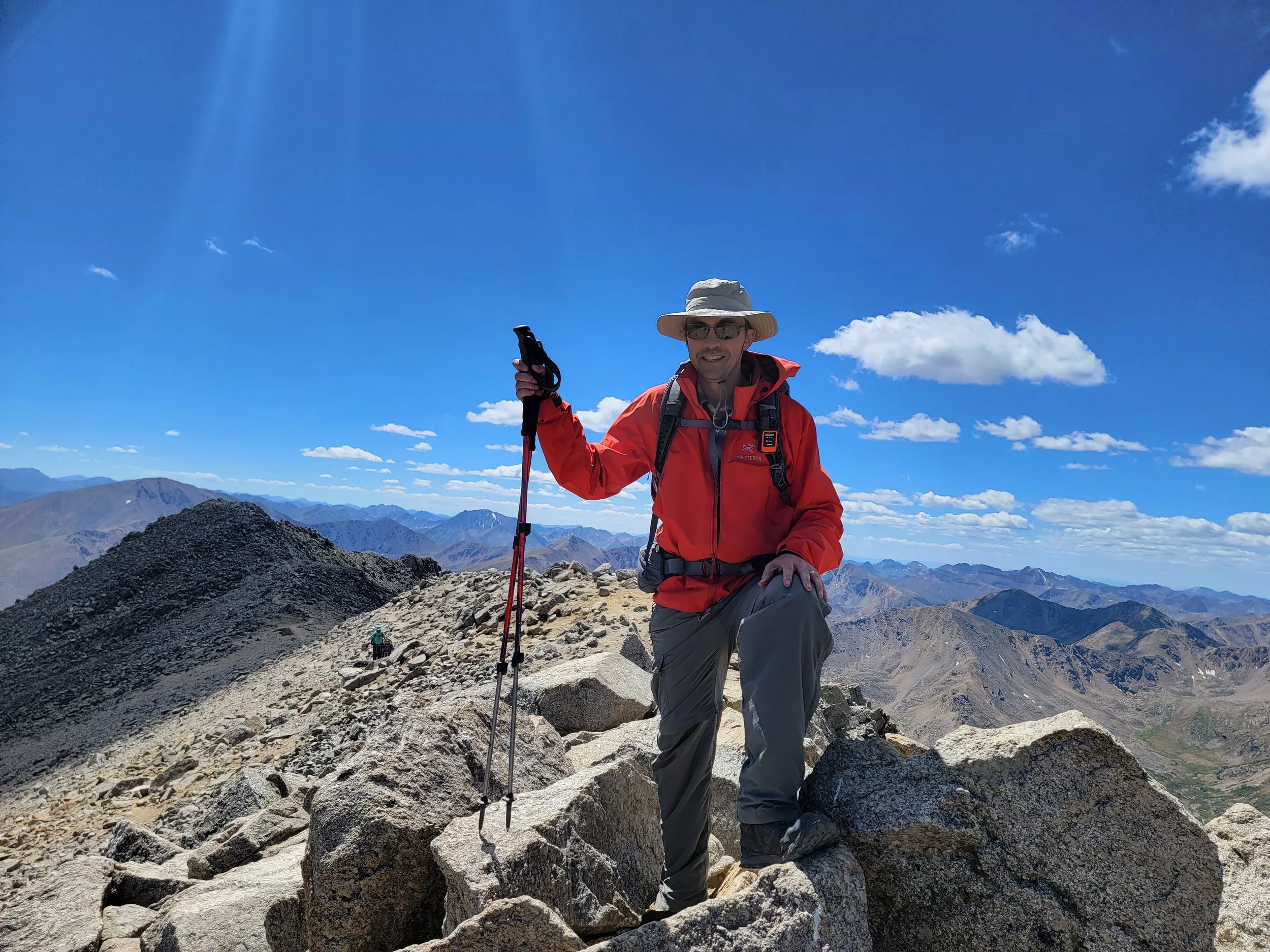 A man wearing outdoor hiking gear, sunglasses, and a broad-brimmed hat, kneeling on a rocky mountain trail under a clear blue sky with scattered clouds, holding hiking poles, with mountain peaks in the background.