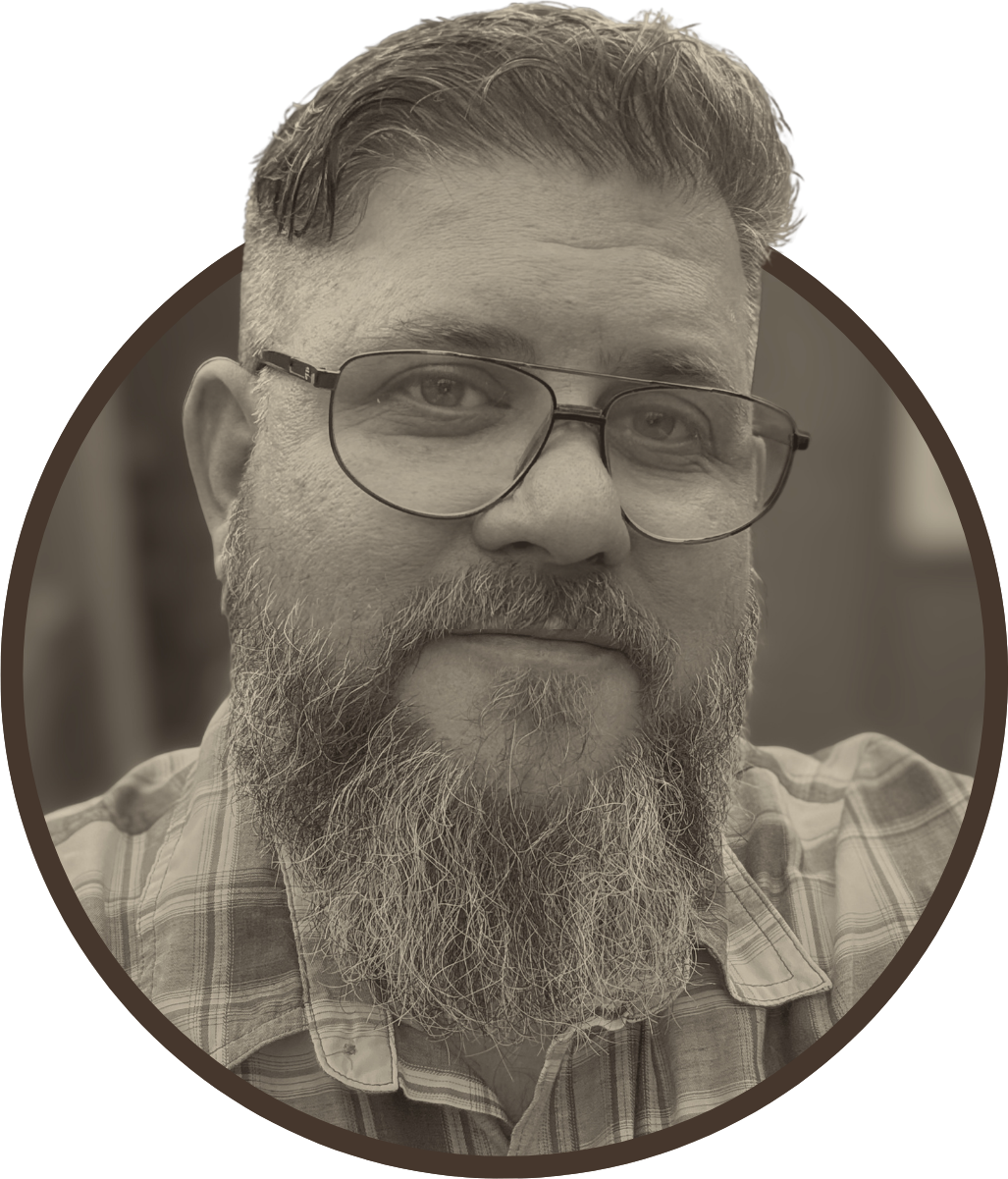 A close-up of a middle-aged man with a full beard, glasses, and short, styled hair, wearing a plaid shirt.
