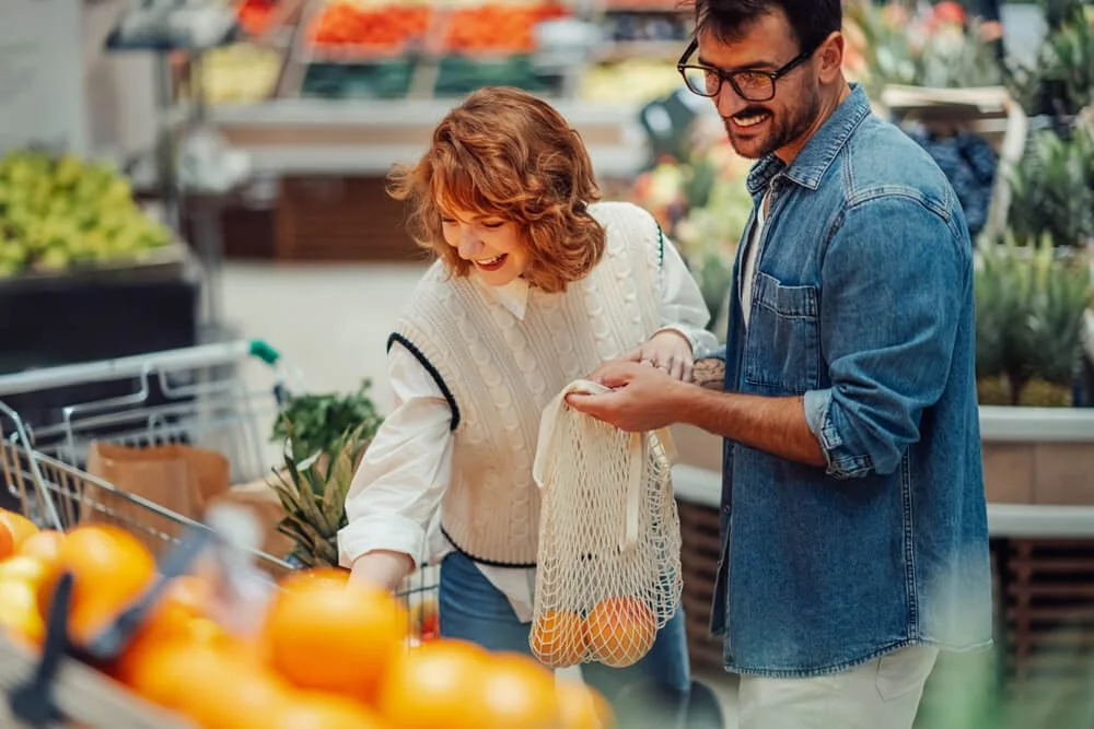 A man and a woman shopping for produce at a grocery store, smiling and holding a mesh bag with oranges.