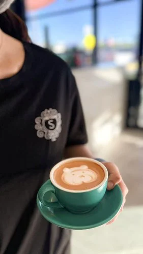 Person holding a green coffee cup with latte art, seated in a cafe with large windows and outdoor view
