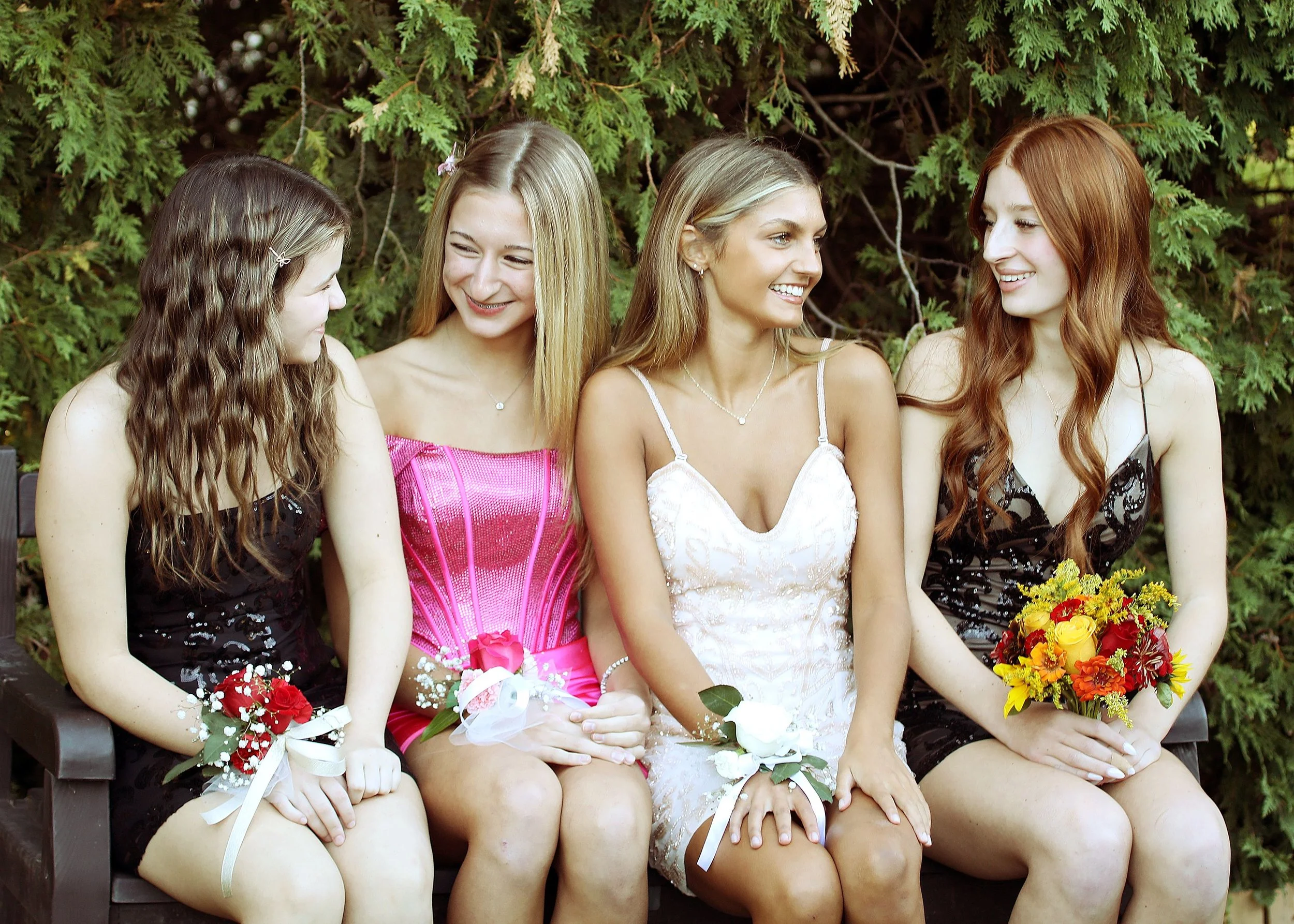Five young women sitting on a park bench surrounded by greenery, wearing dresses, holding flower bouquets, and smiling at each other.