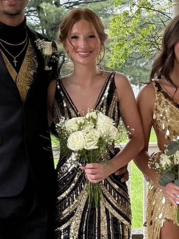 Young woman in a black and gold sequined dress smiling, holding a bouquet of white roses, standing outdoors.