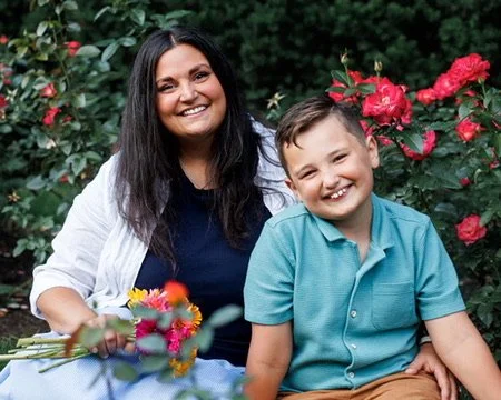 A woman and a young boy sitting outdoors in front of pink and red flowers, smiling at the camera.