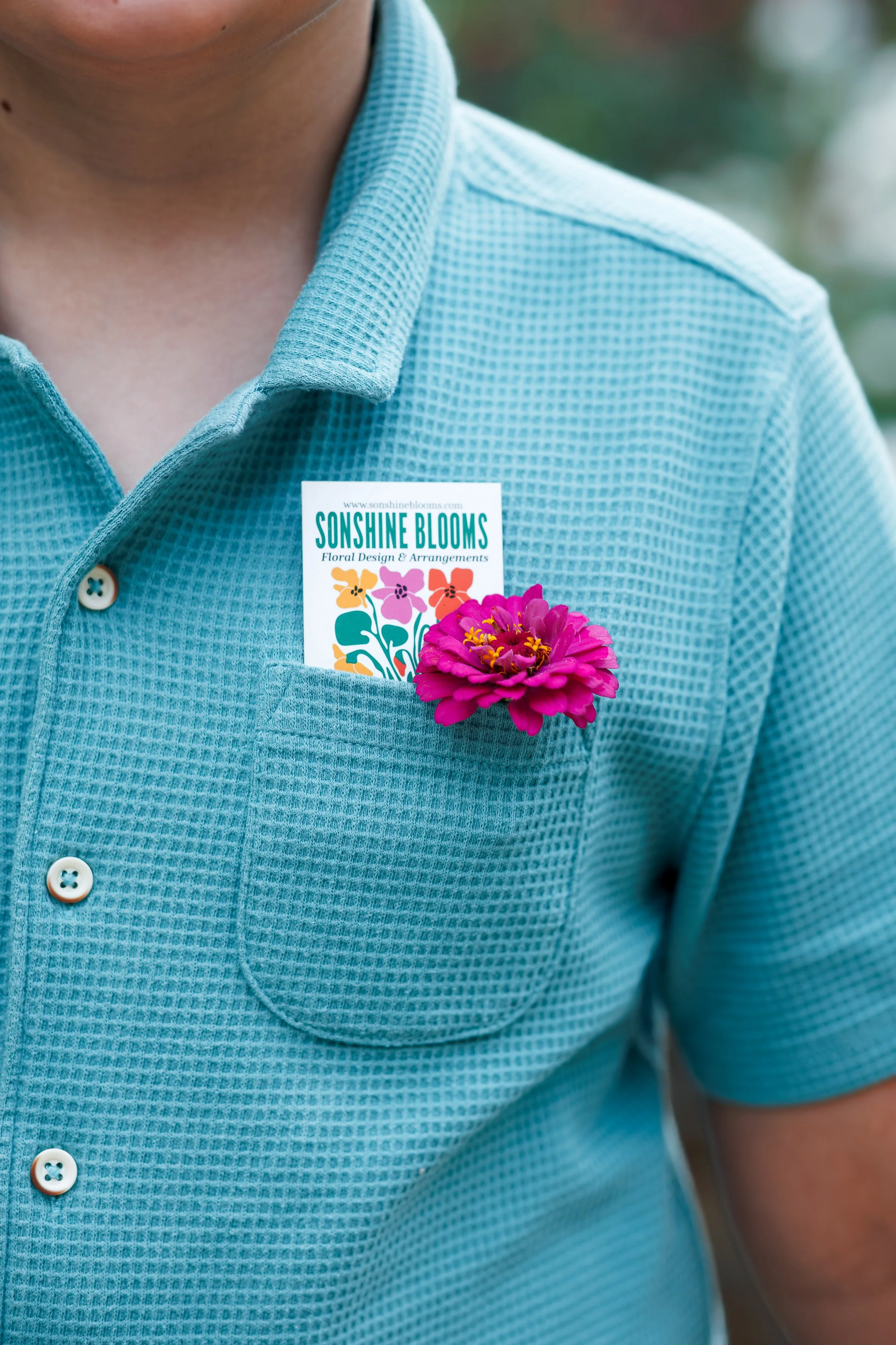 Close-up of a person wearing a blue, textured button-up shirt with a pink flower pinned to the chest pocket and a business card from Sonshine Blooms featuring colorful flowers, floral design, and website URL.
