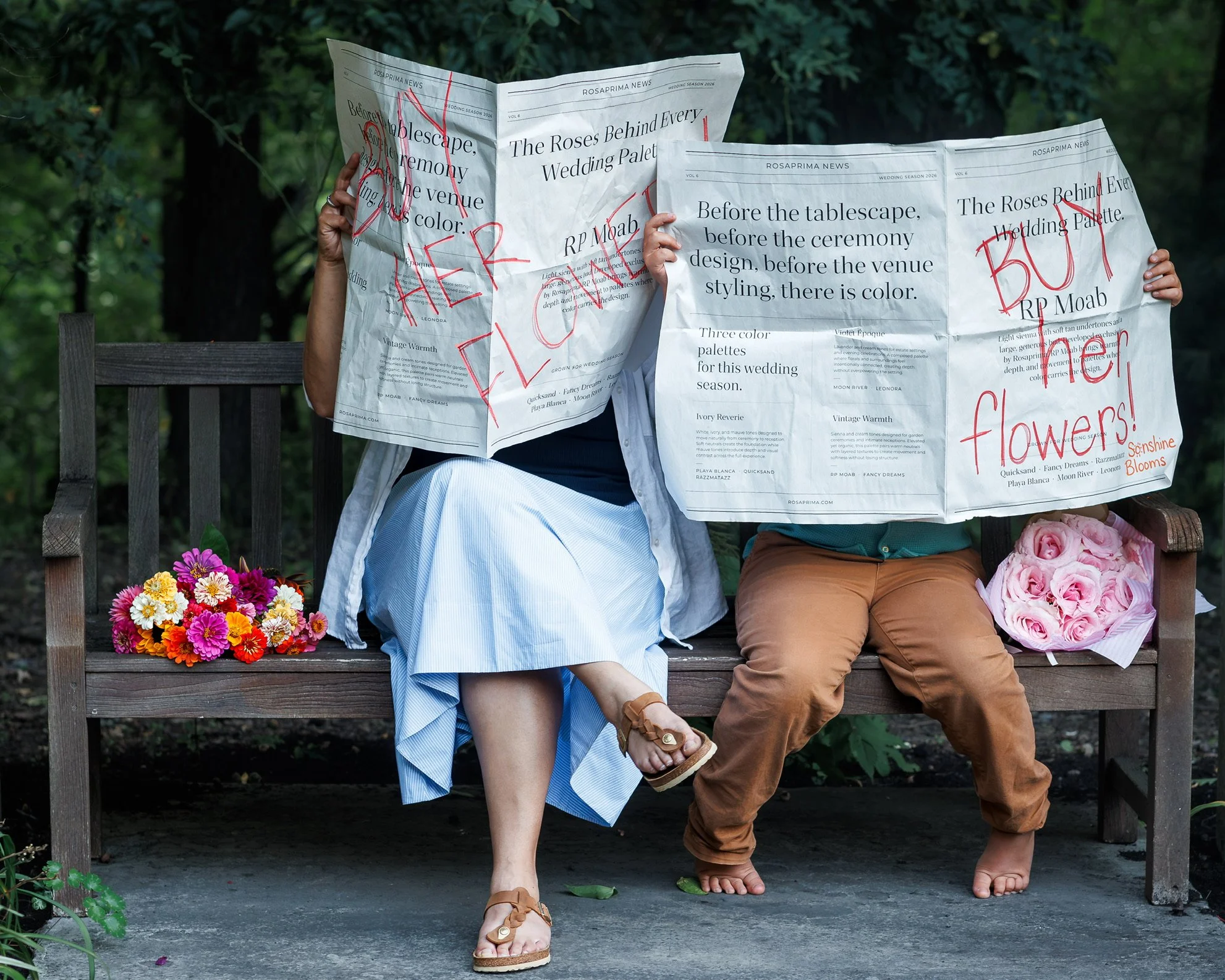 Two people sitting on a park bench, each holding newspapers with red handwriting on them. The first person's newspaper reads 'BETTER FLOWERS' and the second reads 'BLOOMS'. The person on the left is wearing a blue and white dress with sandals, and the person on the right is wearing brown pants and is barefoot. There are bouquets of flowers at their feet, and greenery surrounds the bench.
