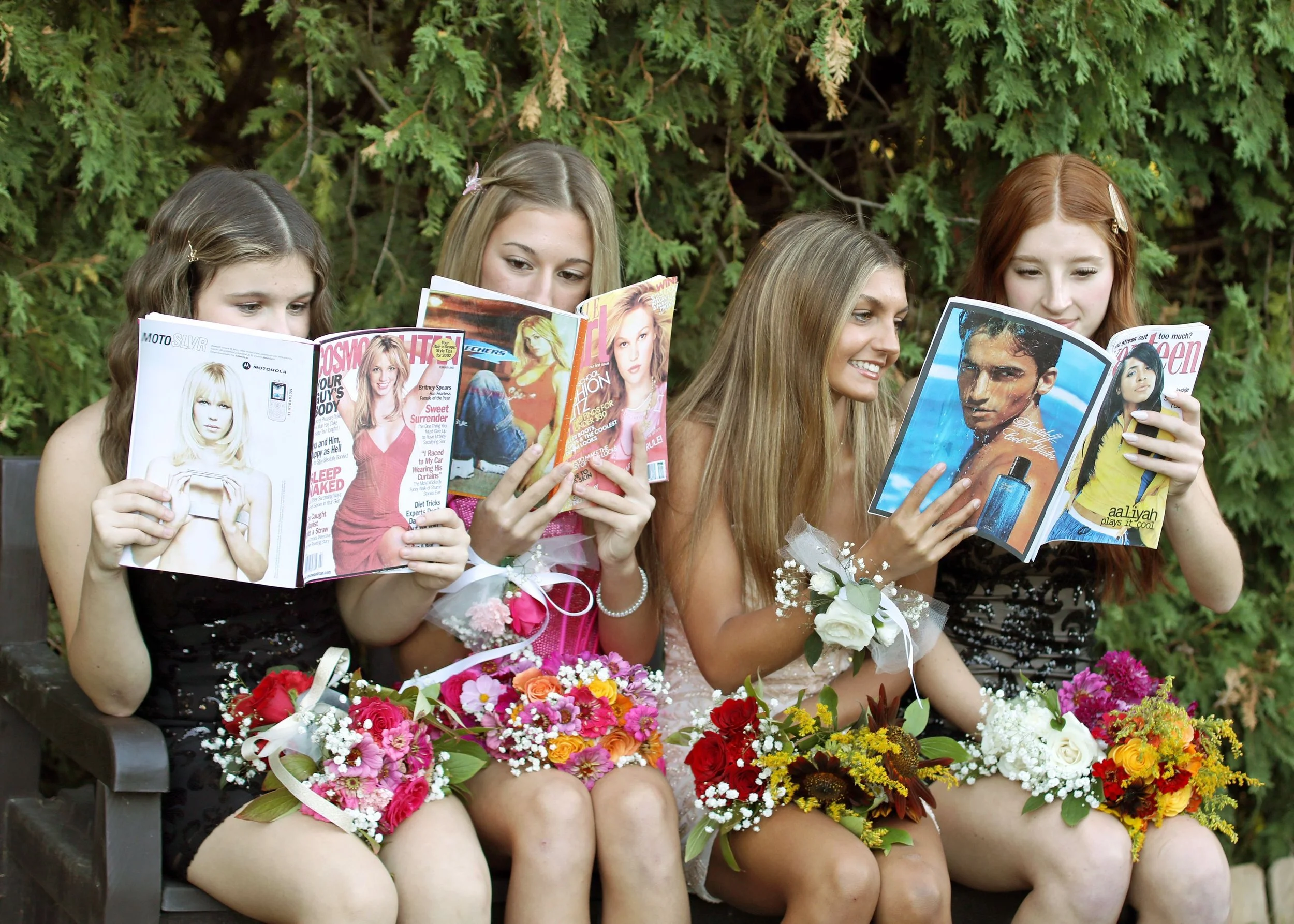 Four young women sitting outdoors reading magazines and holding bouquets of flowers, with a background of green trees.