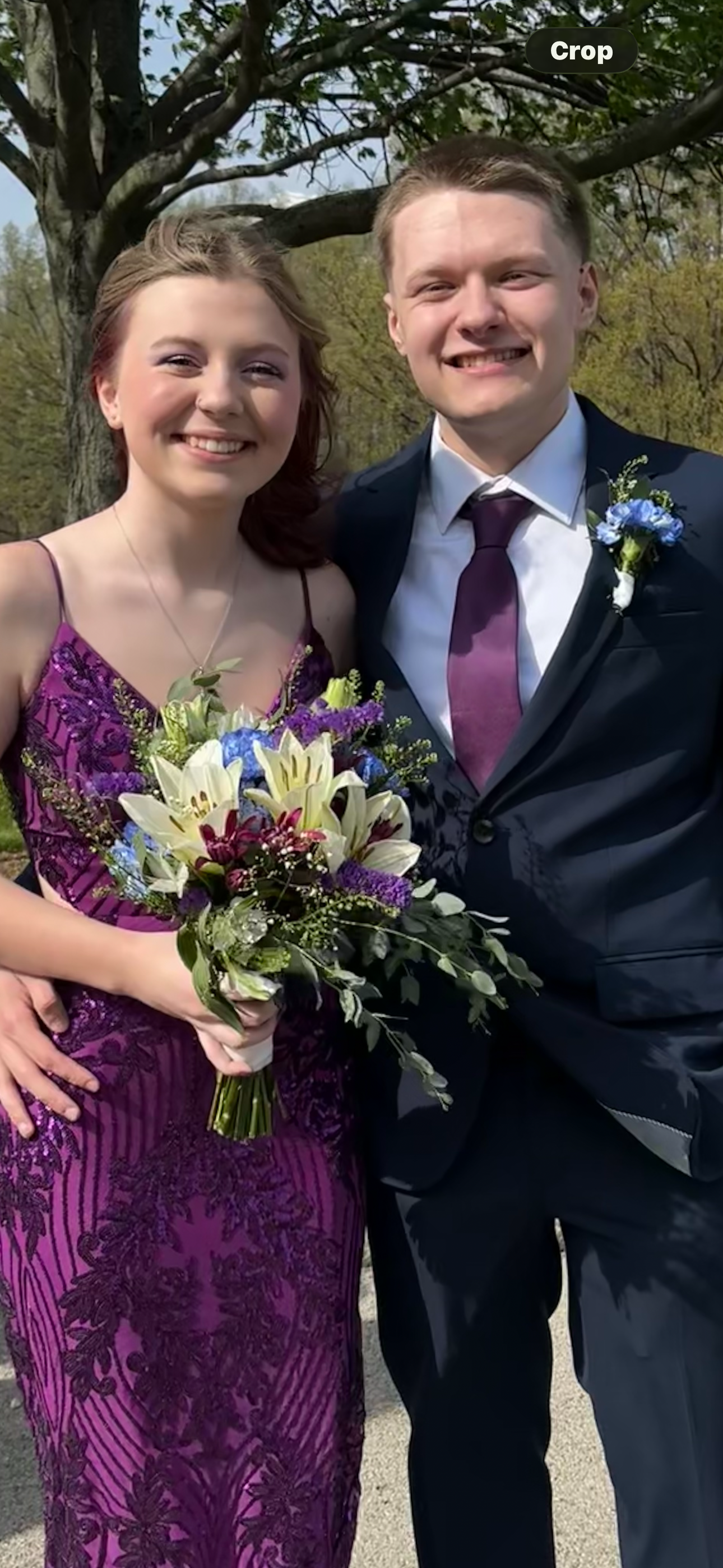 A young woman and a young man in formal attire standing outdoors with trees in the background, smiling, and holding a bouquet of flowers.
