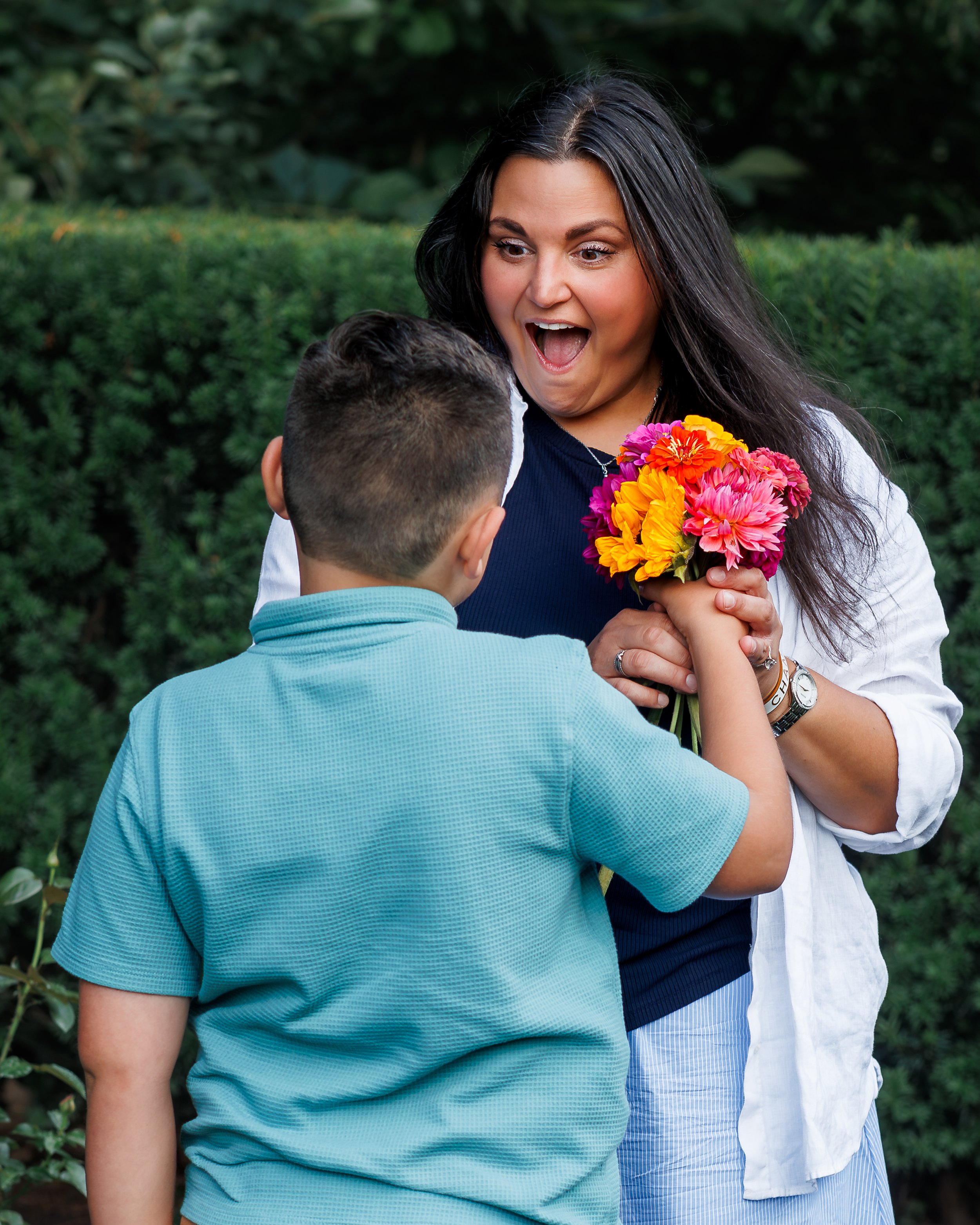 A woman with long dark hair, wearing a white shirt, holds a colorful bouquet of flowers and reacts with surprise, while a young boy with short hair, wearing a light blue polo shirt, stands in front of her in a garden setting.