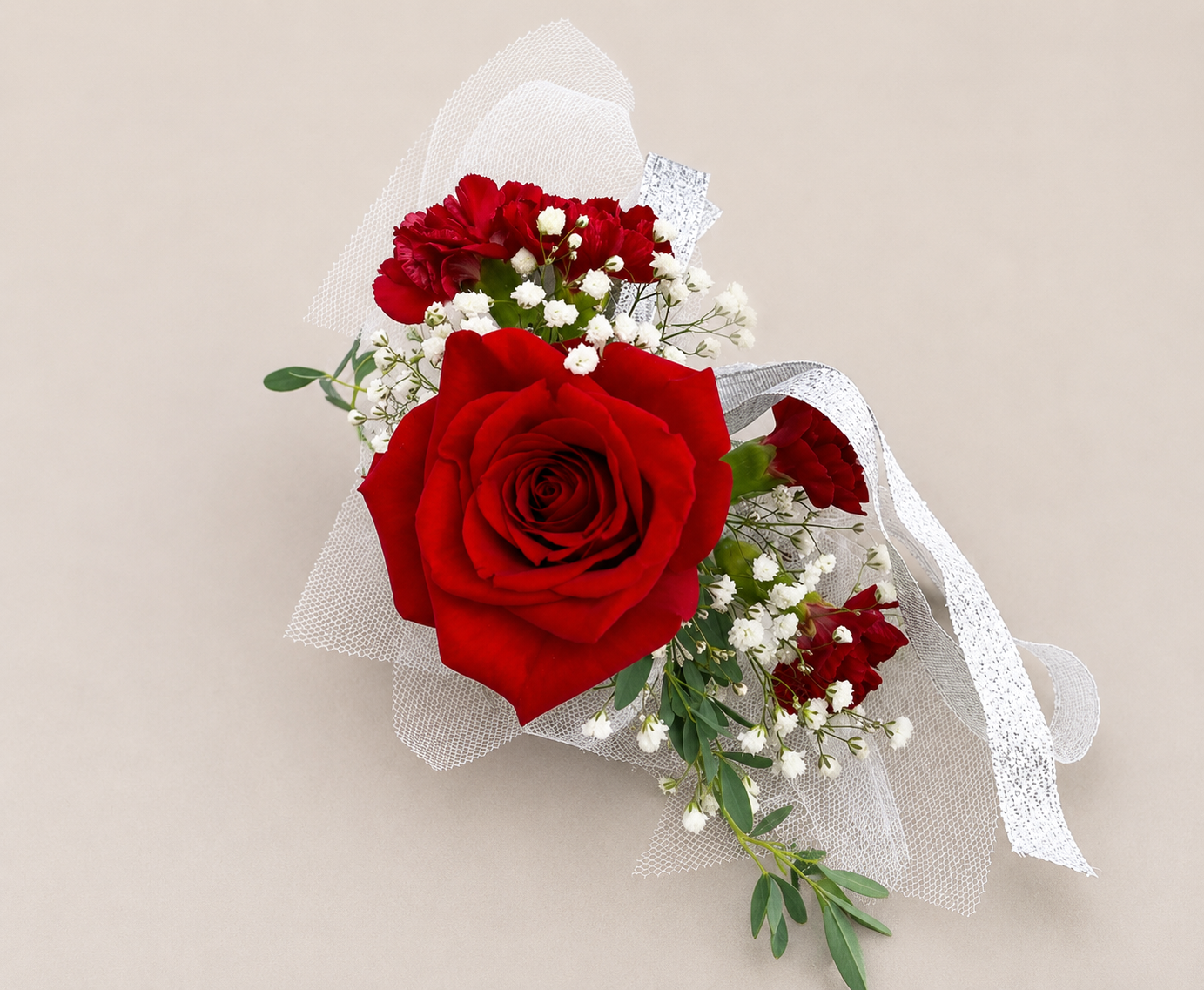 A bouquet of red roses, white baby's breath, and red carnations wrapped in white mesh ribbon on a beige background.