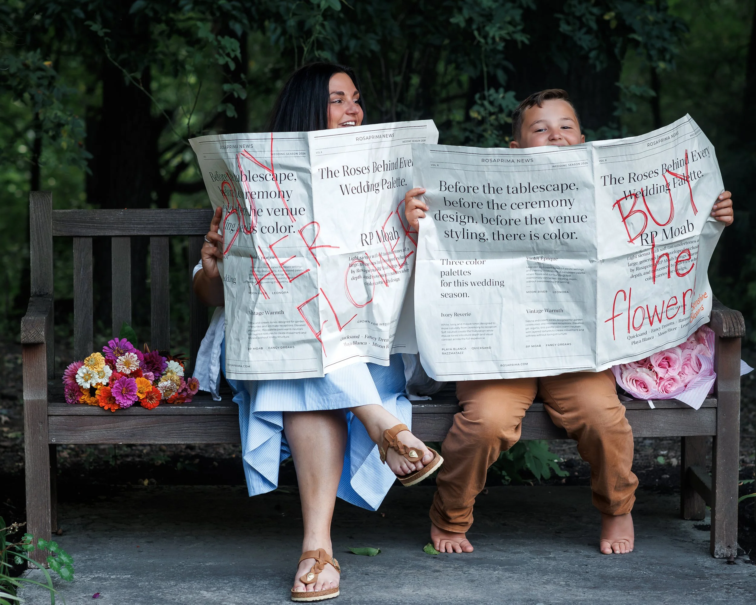 A woman and a young boy sitting on a wooden bench outdoors, holding large sheets of newspaper in front of their faces. The woman has dark hair and is smiling, wearing a blue and white striped dress. The boy is wearing a light purple shirt and brown p
