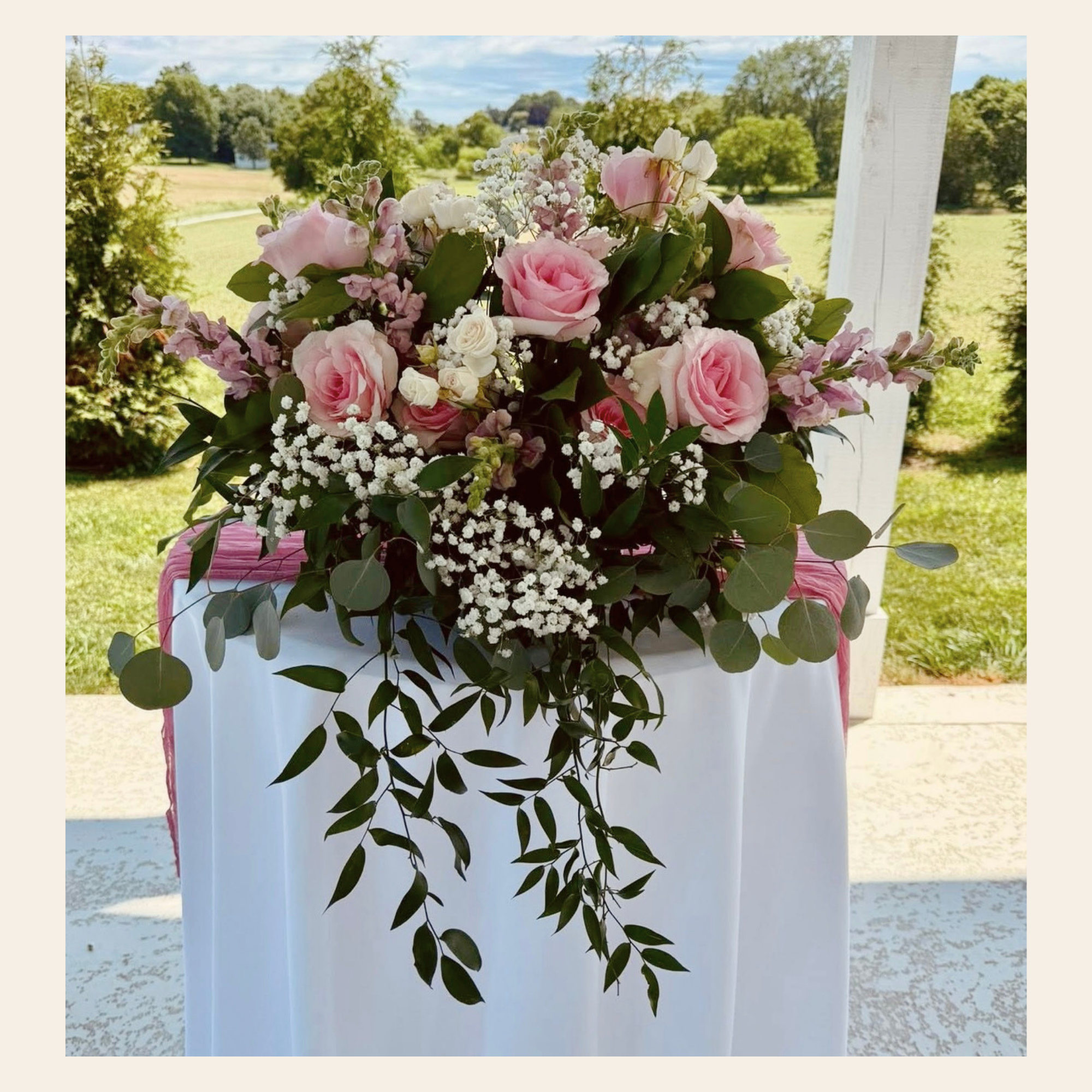 A floral arrangement with pink roses, white spray roses, baby's breath, and greenery, placed on a white and pink decorated stand outdoors during daytime.