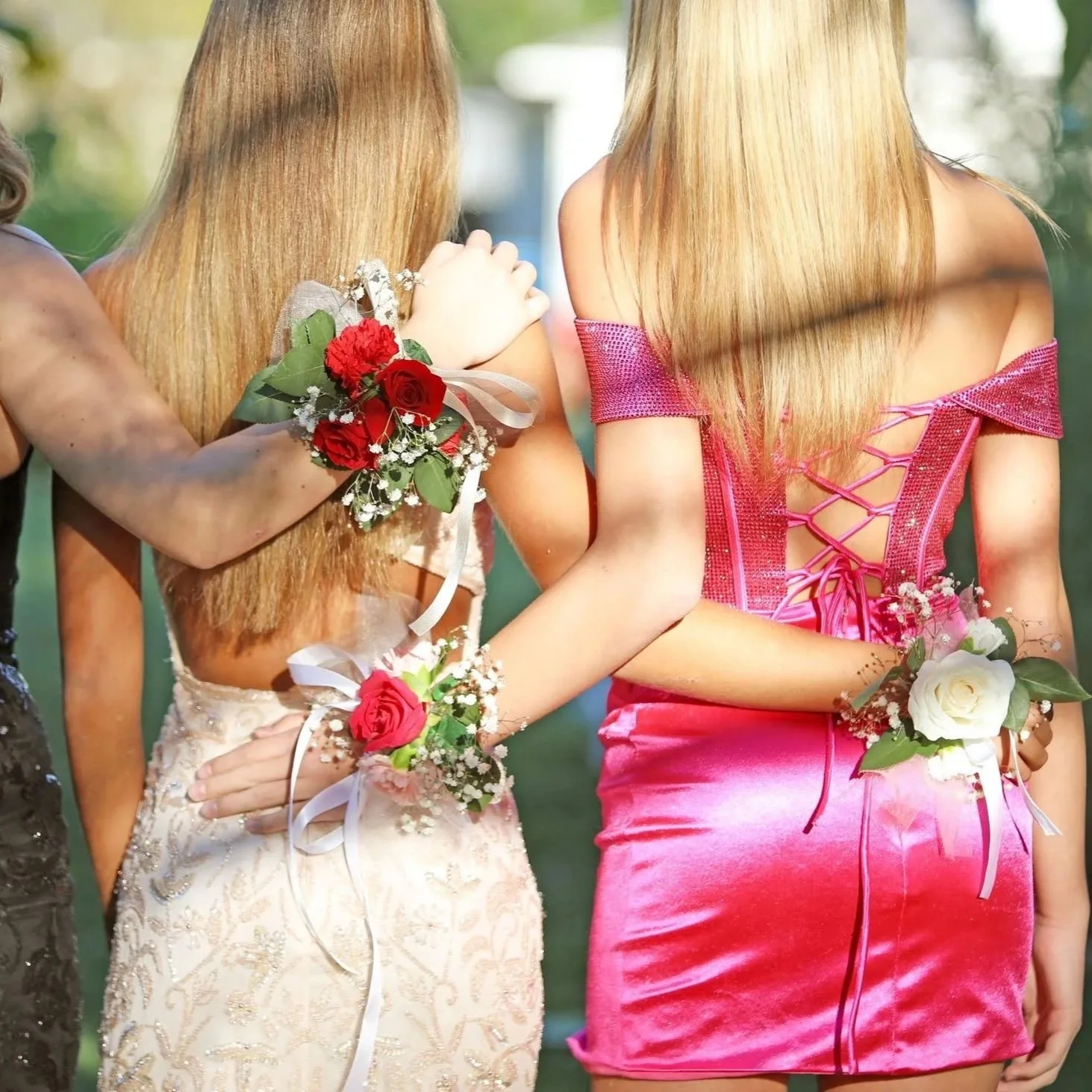 Two women with long blonde hair holding hands at a formal event, wearing floral corsages and dresses, outdoors with blurry greenery in the background.