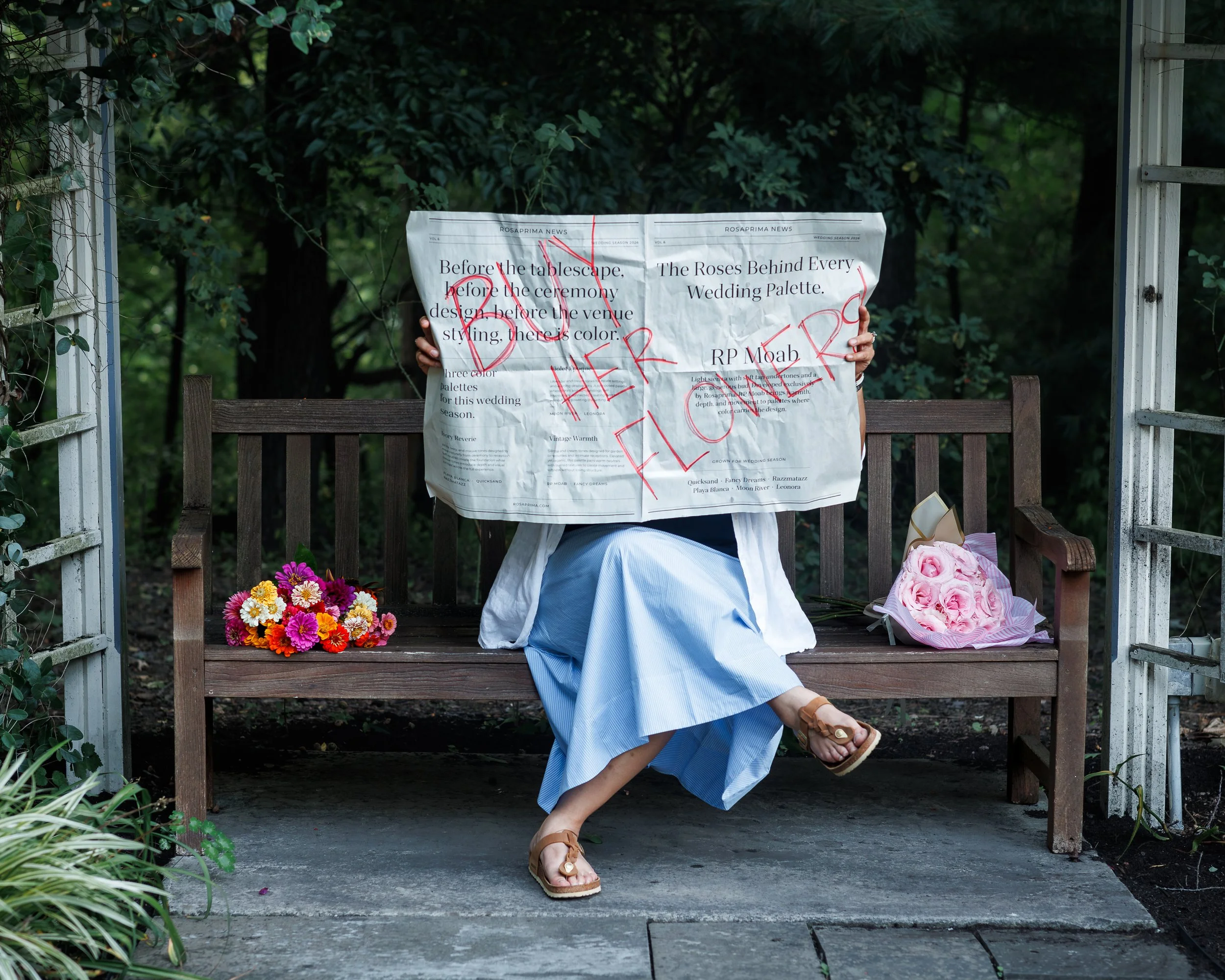 Person sitting on a wooden bench outdoors holding a large newspaper in front of their face, with flowers on the ground and greenery in the background.