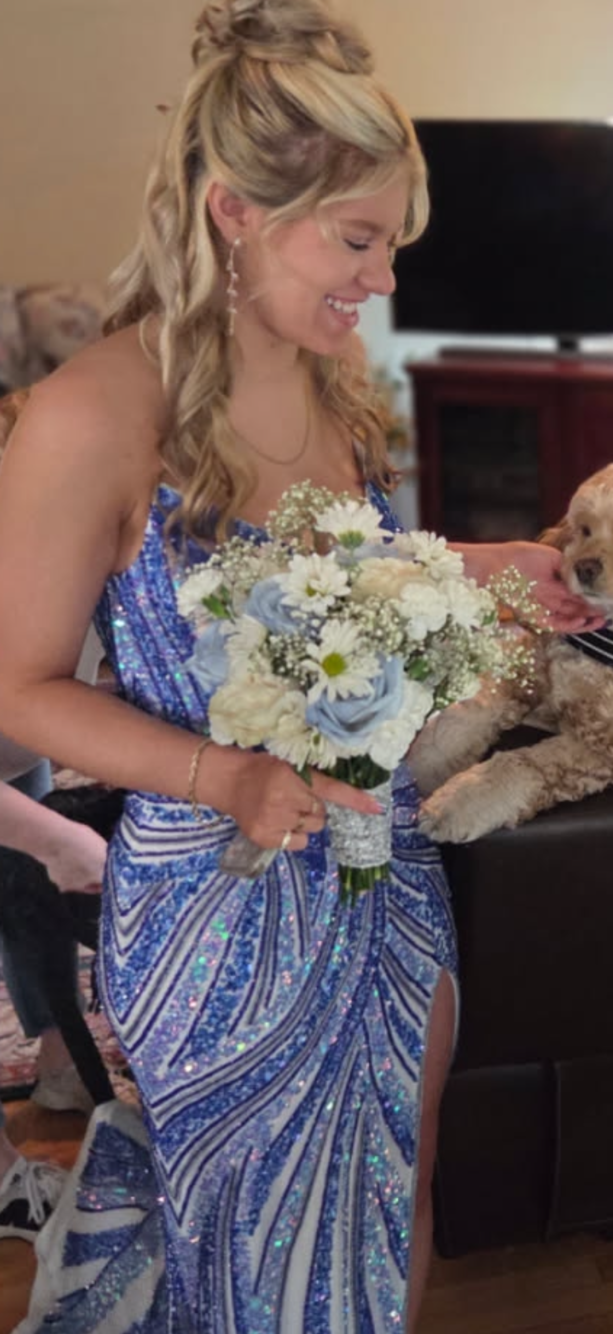 A woman in a sparkly blue dress holding a bouquet of white and blue flowers, with a dog nearby, in an indoor setting.